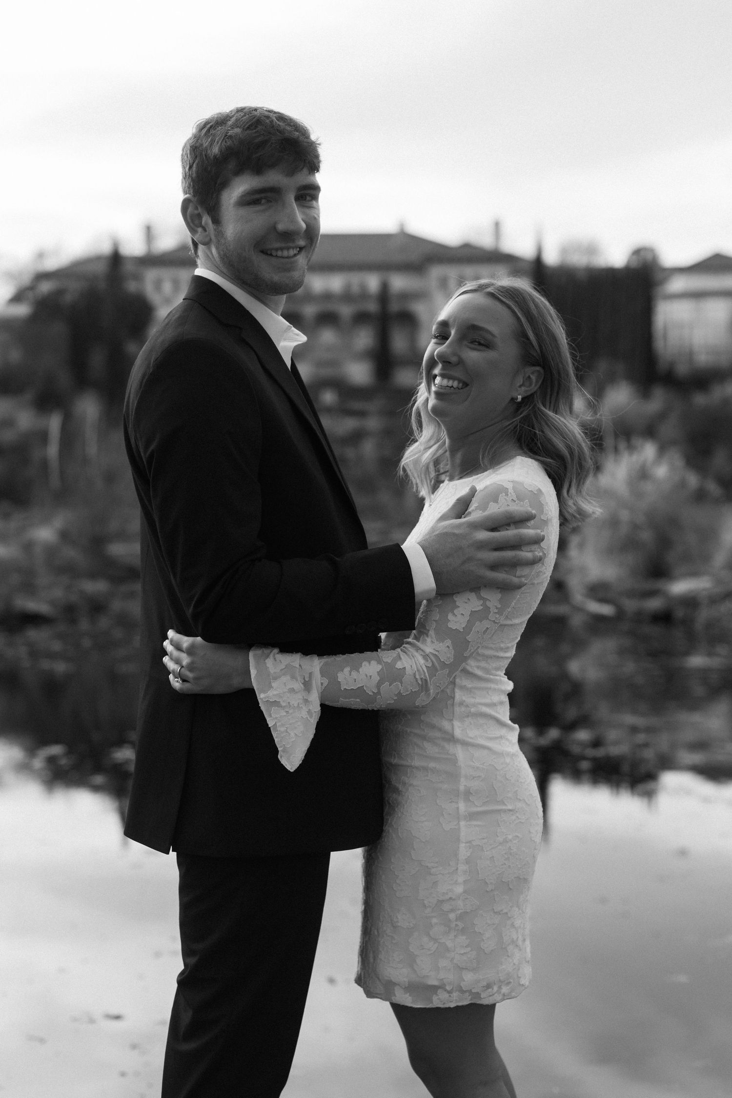 A couple in formal wear shares a joyful moment by a reflective pond.