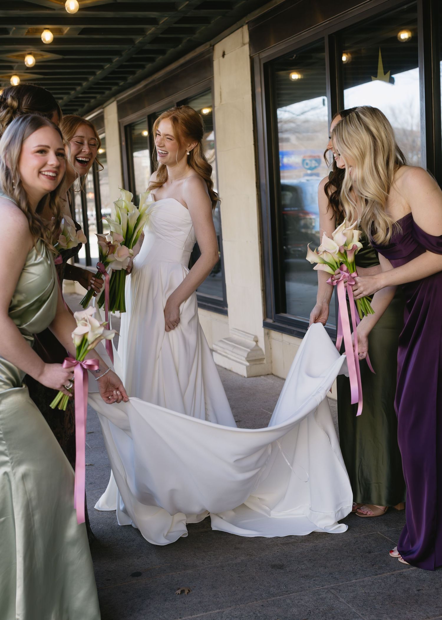 Bridesmaids in purple and sage dresses help arrange a bride's wedding gown.