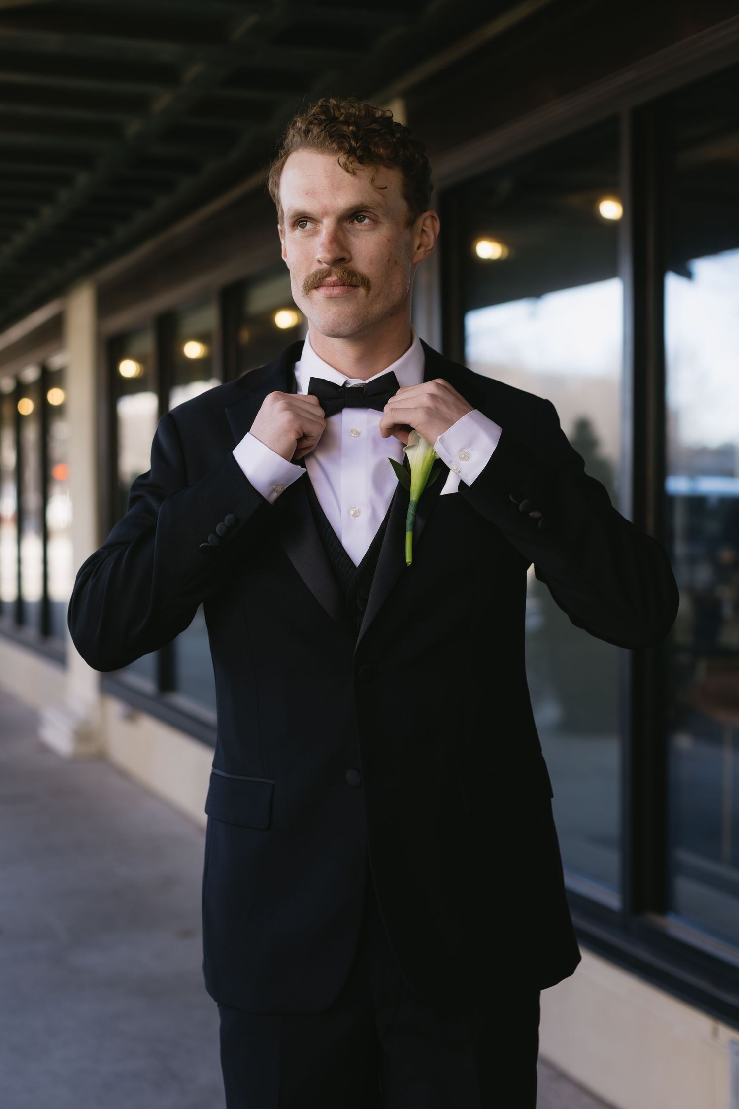 Groom adjusts his bowtie while wearing classic black tuxedo in an industrial setting.