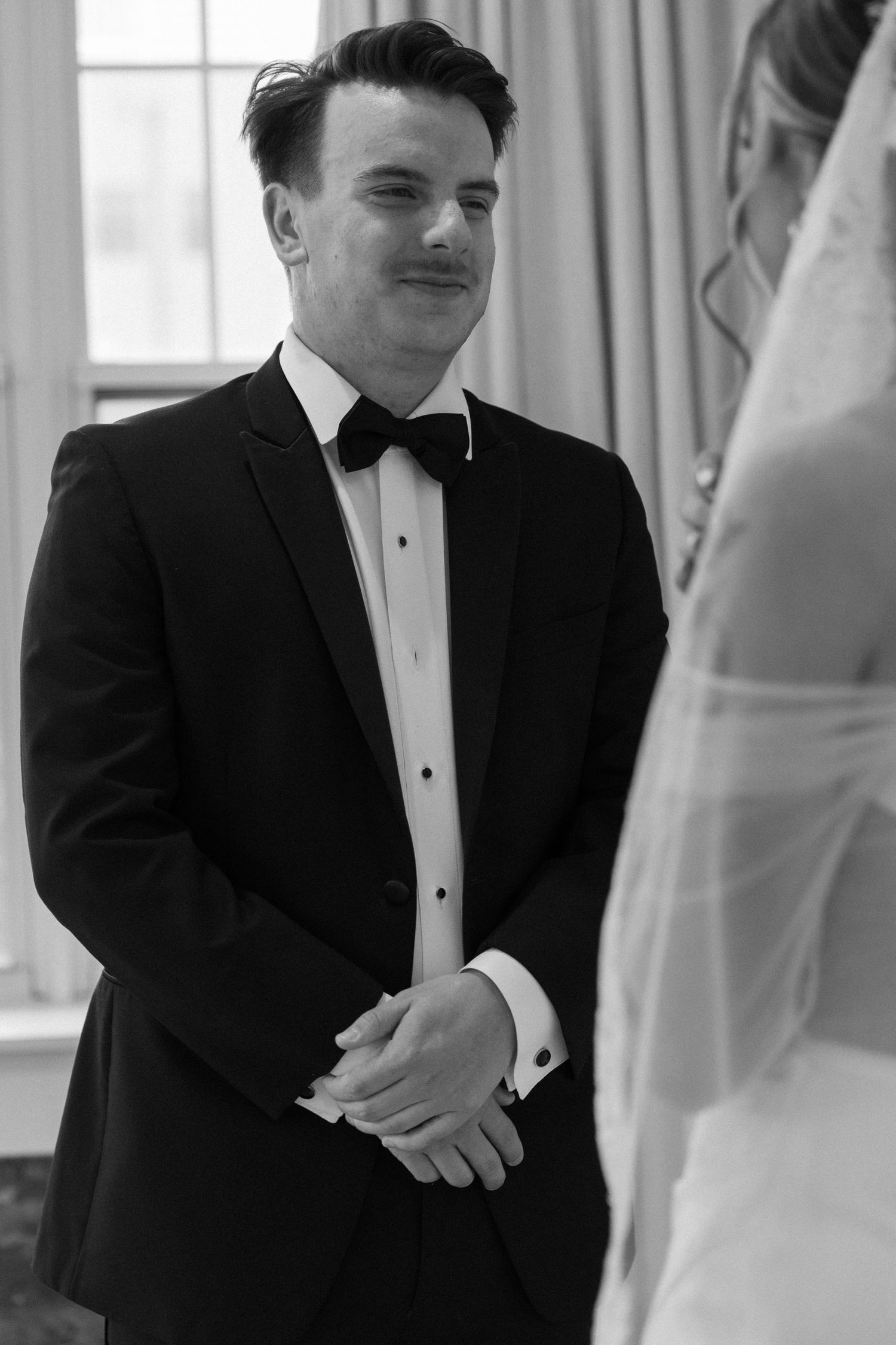 A formal black and white portrait of a groom in a classic tuxedo during the wedding ceremony.