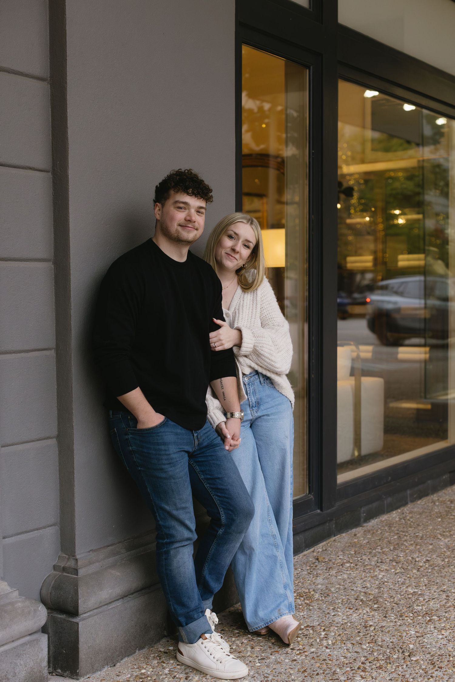 A couple leaning against a storefront wall wearing casual outfits of jeans and sweaters.