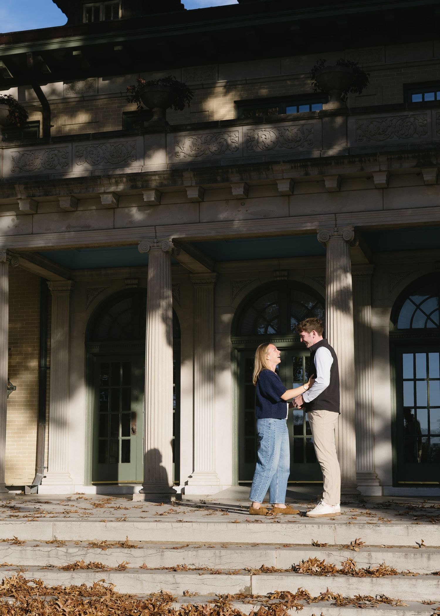 A couple poses together against a stone building with columns and arched windows.