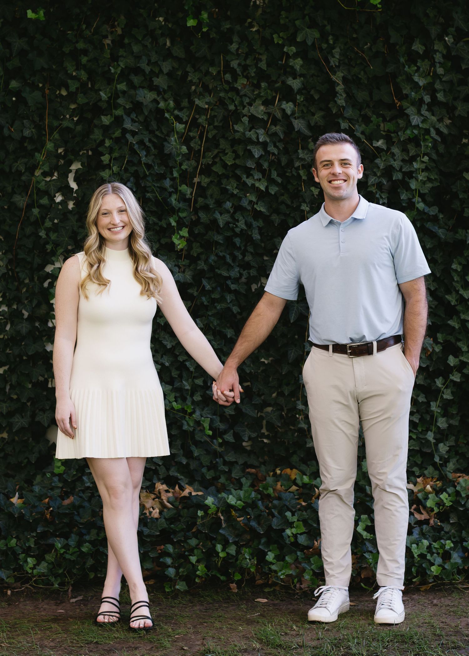A couple poses against an ivy wall backdrop in matching light-colored outfits.