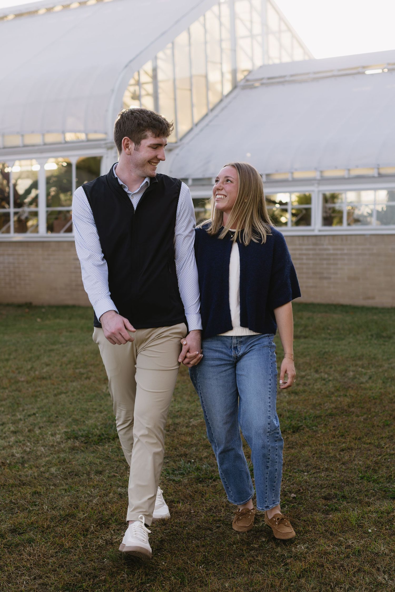 A couple walking together on grass wearing casual outfits of jeans, khakis and dark vests.