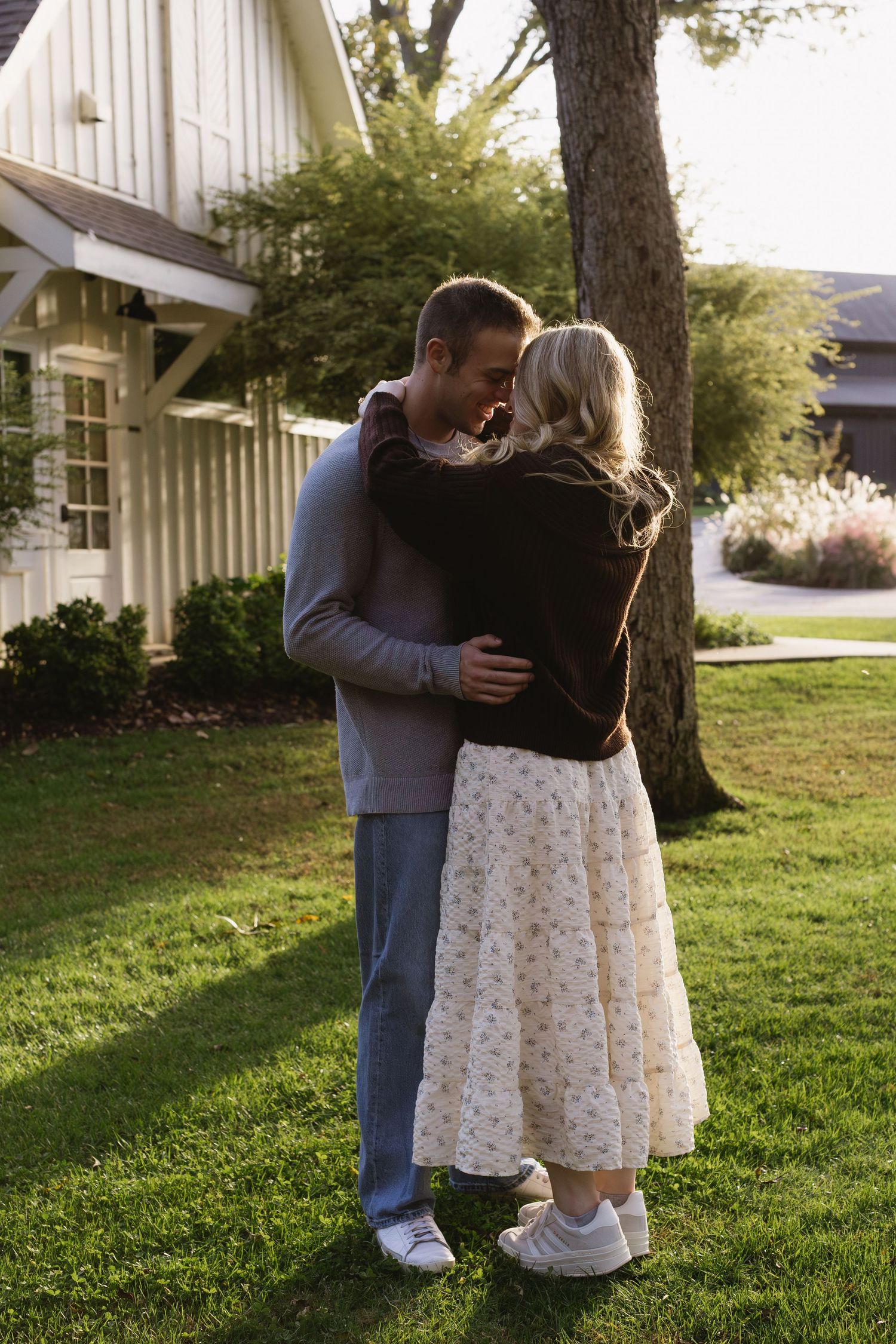 A couple embraces near a white building with trees and grass in the background.