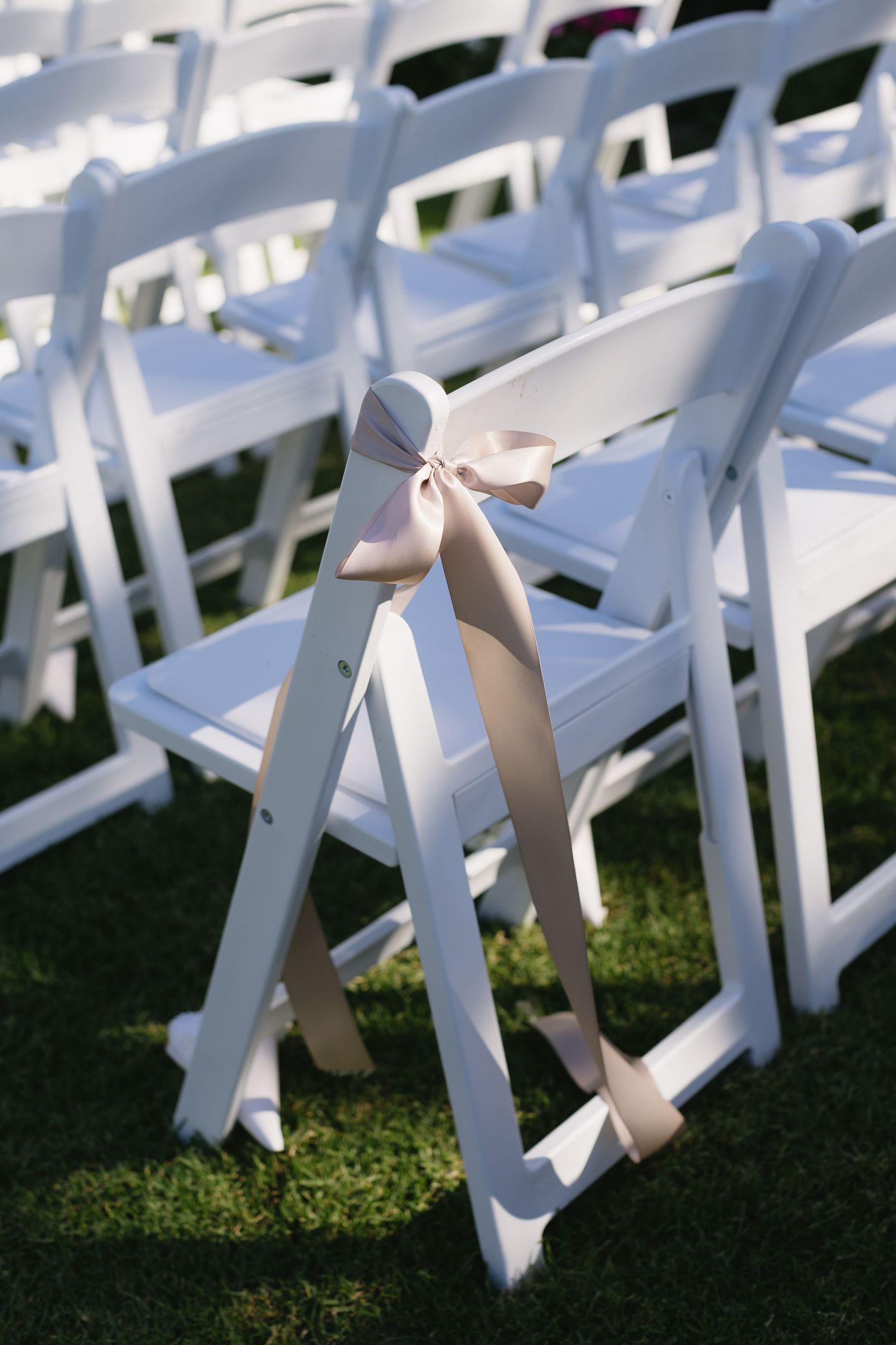 White wedding ceremony chairs with blush pink ribbons arranged on a sunny lawn.