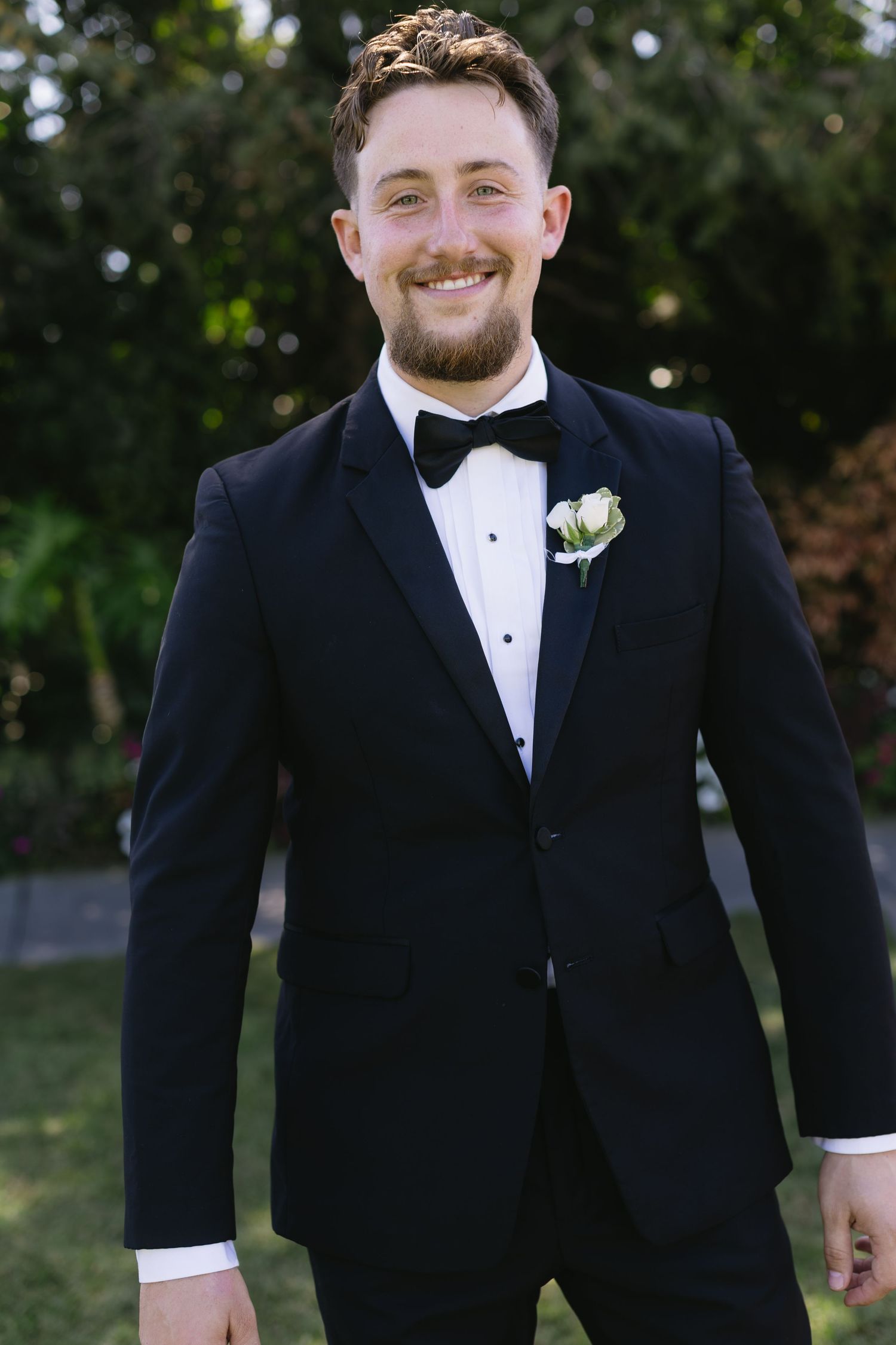Groom in classic black tuxedo and white bowtie smiles warmly during outdoor wedding ceremony.