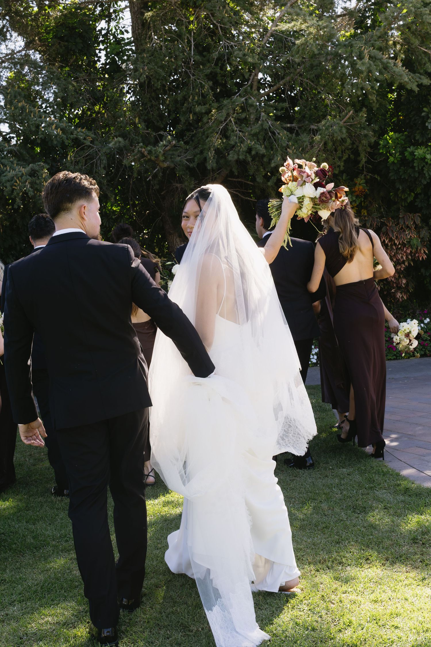 Wedding party members walk together on garden path during outdoor celebration.