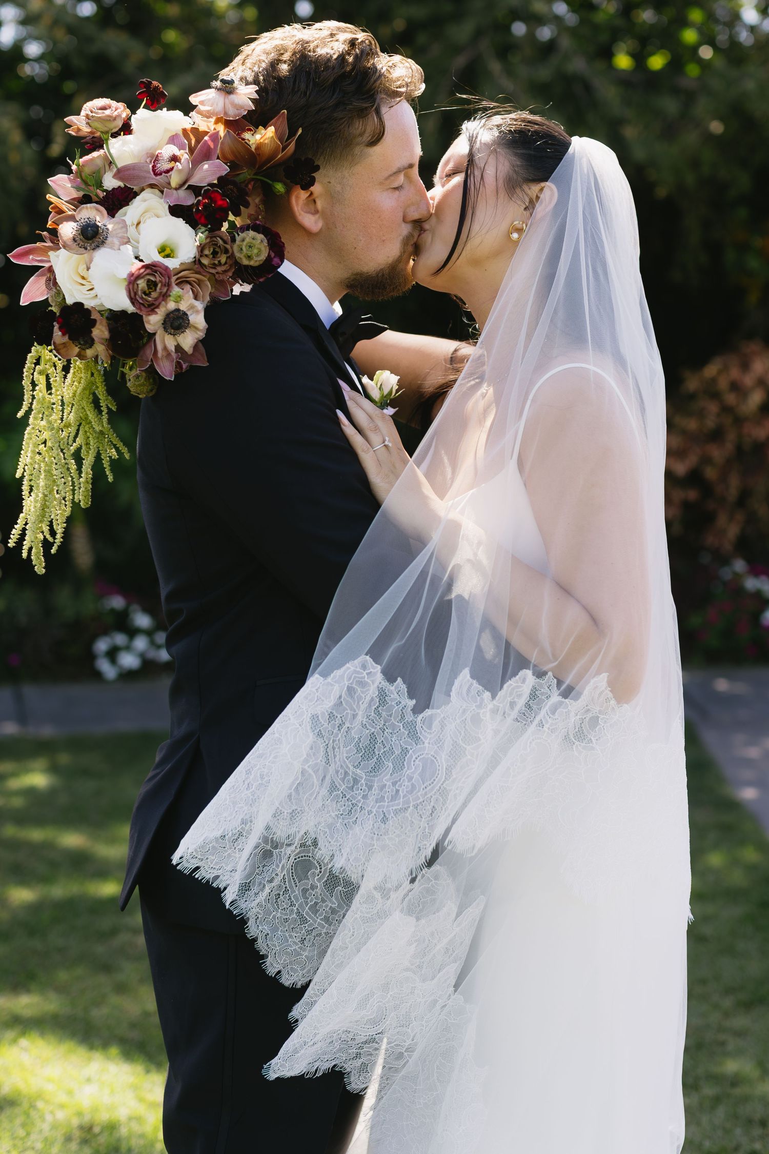 Wedding couple embracing under a lace veil with floral arrangements in outdoor setting.