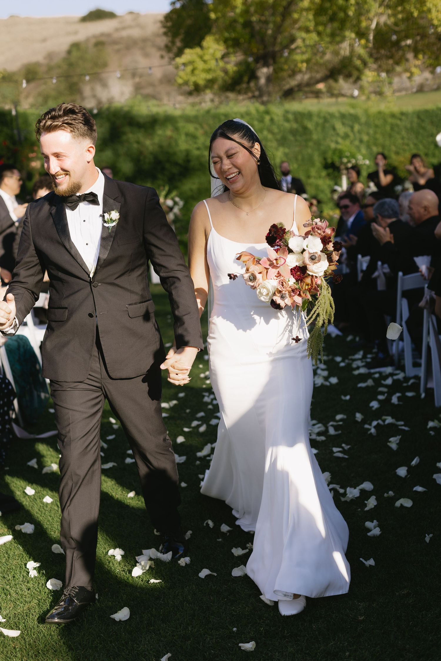 Couple walking down aisle with rose petals scattered on ground at outdoor wedding.