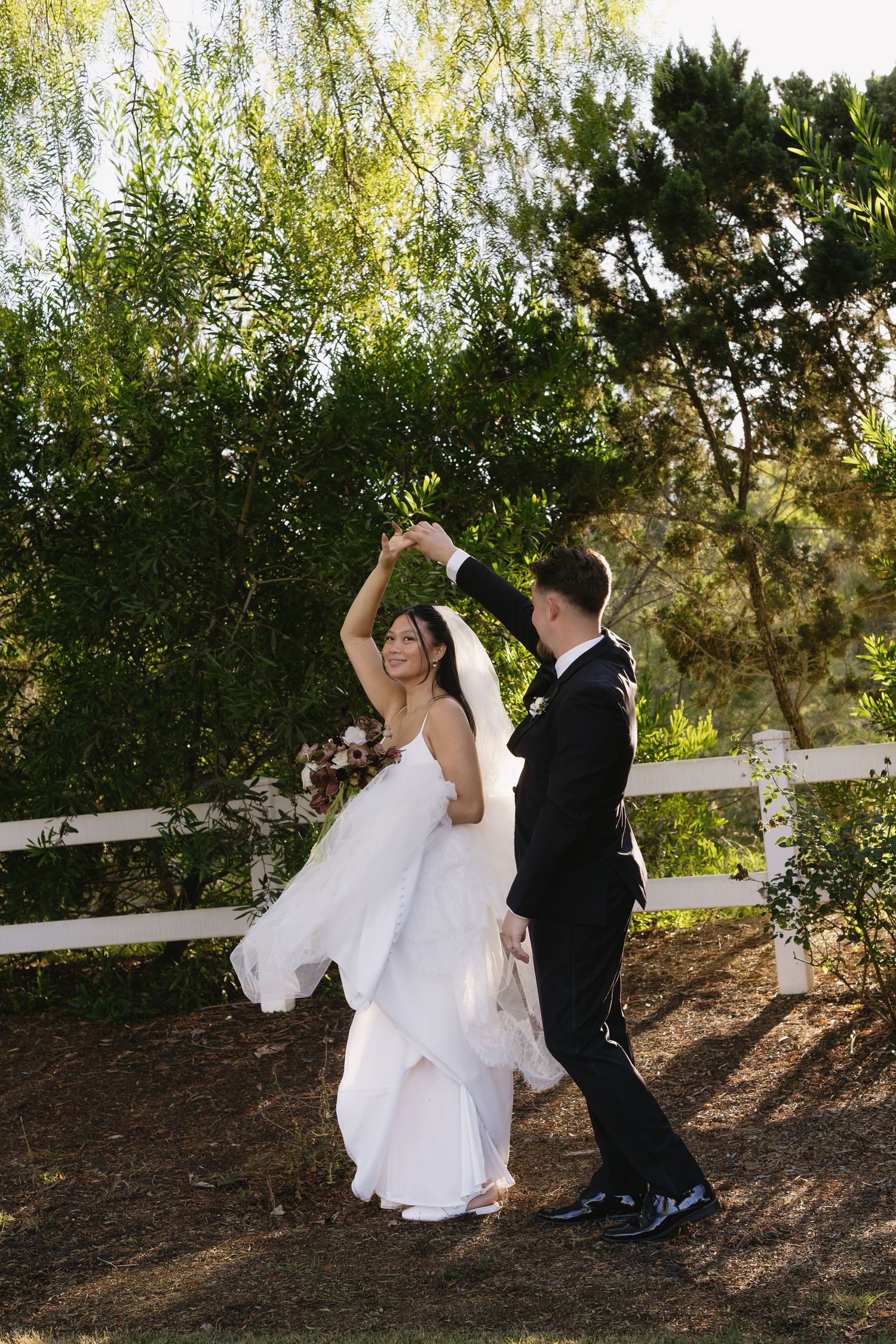 A playful wedding couple dances together near a white fence with trees in the background.