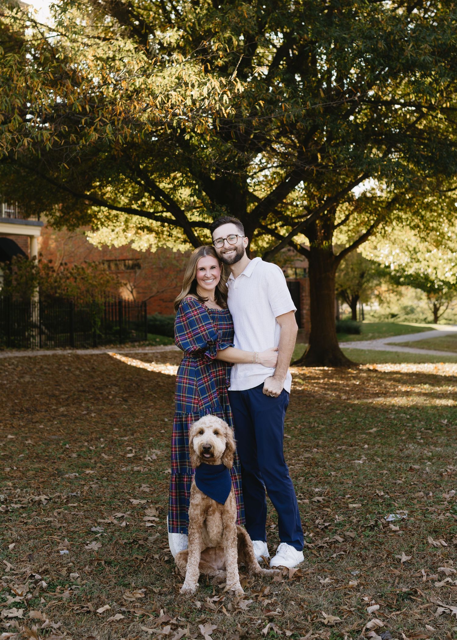 A couple and their goldendoodle dog pose together for autumn photos on fallen leaves in a park setting.