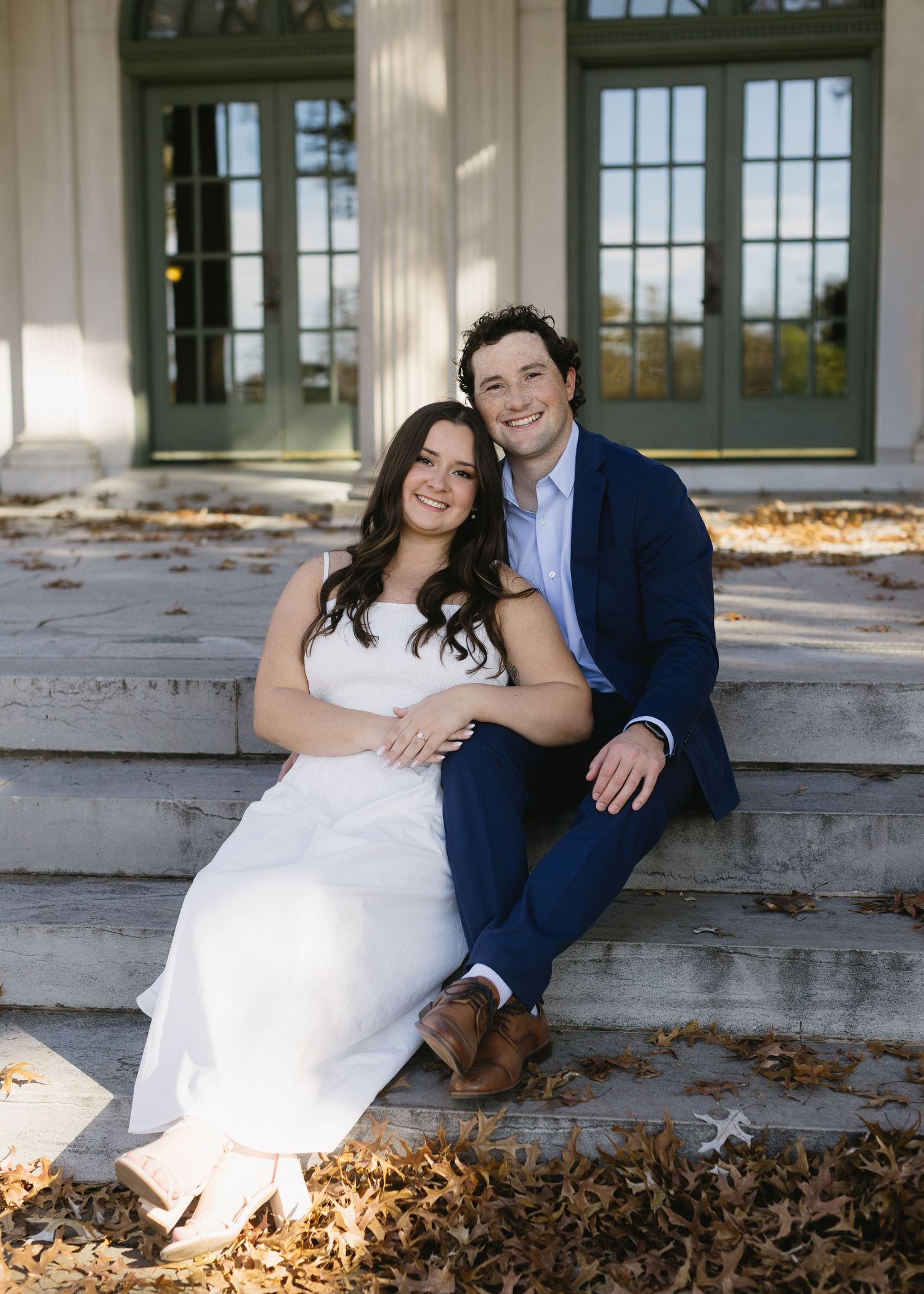 A couple in wedding attire sits on stone steps surrounded by autumn leaves in a romantic outdoor photo series.