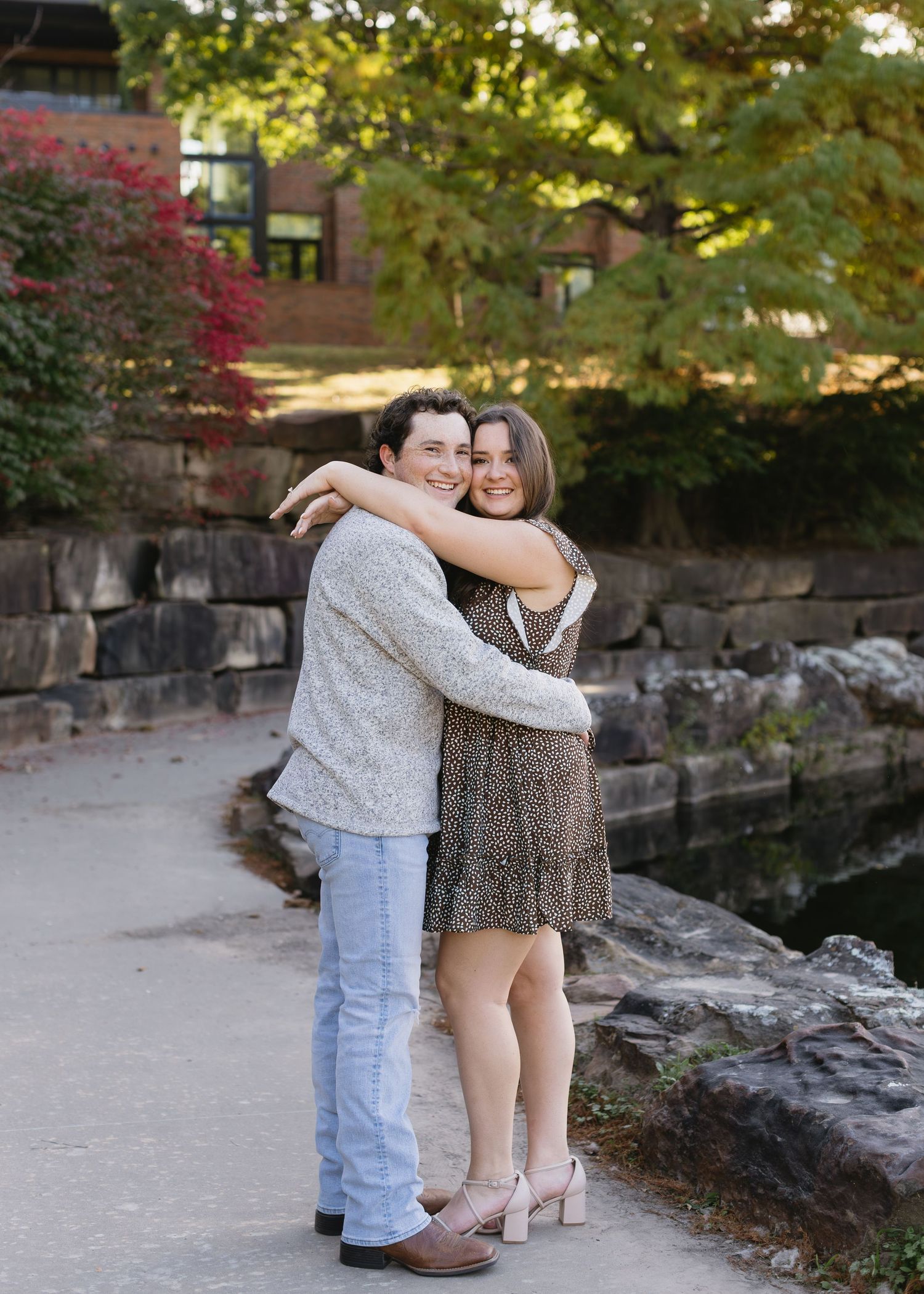 Two young children embrace while standing on a rocky path surrounded by autumn foliage and stone walls.