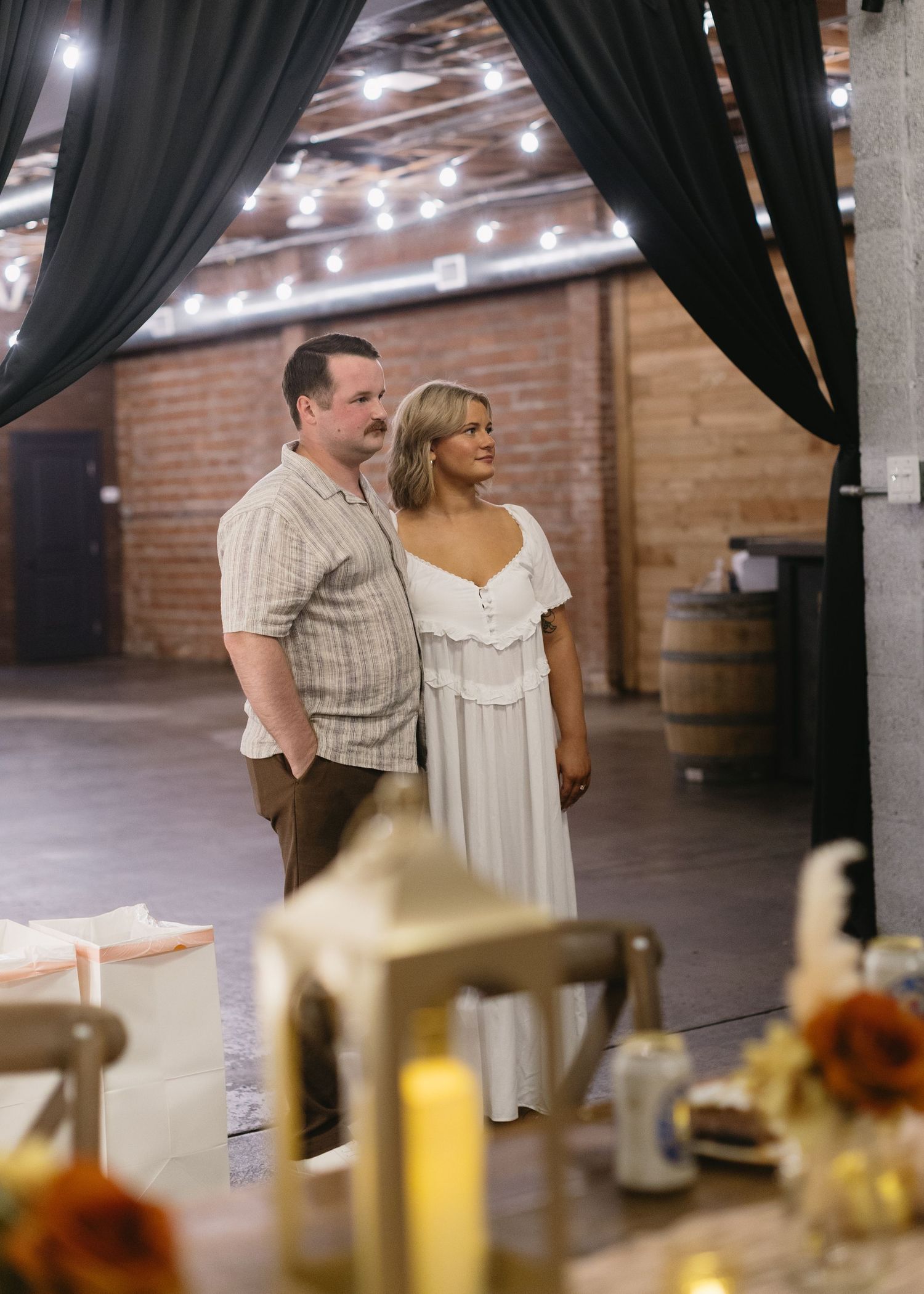 Wedding couple standing in rustic venue with string lights and dark curtains.