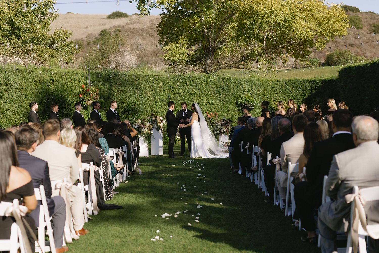Outdoor garden wedding ceremony with guests seated in white chairs and greenery backdrop in natural lighting.