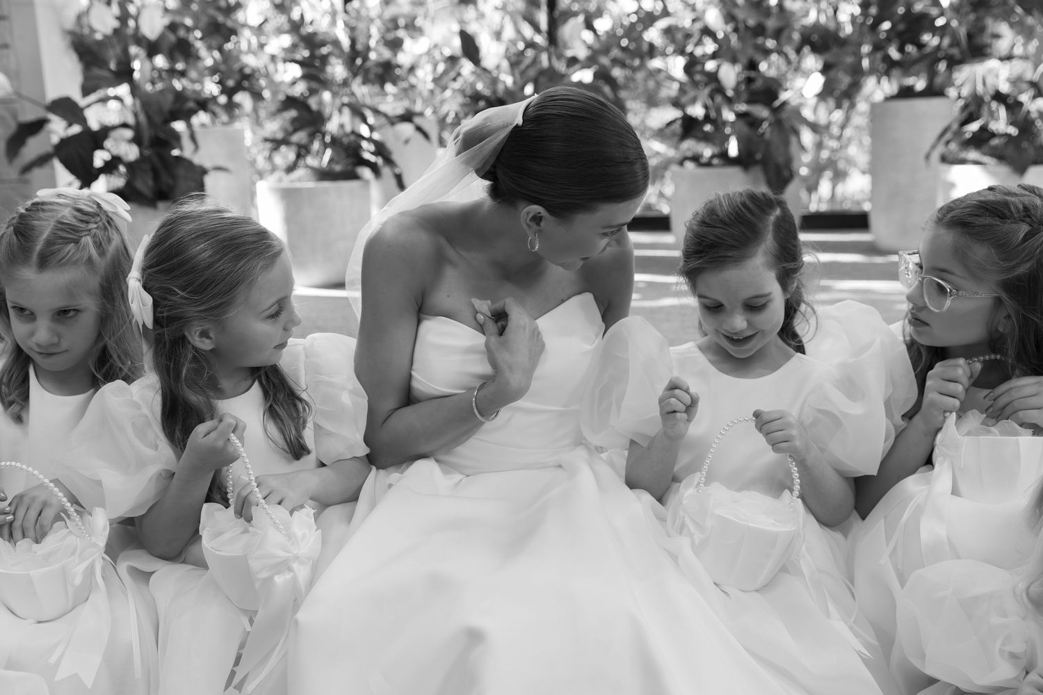 A row of young flower girls in white dresses line up peeking through their hands.