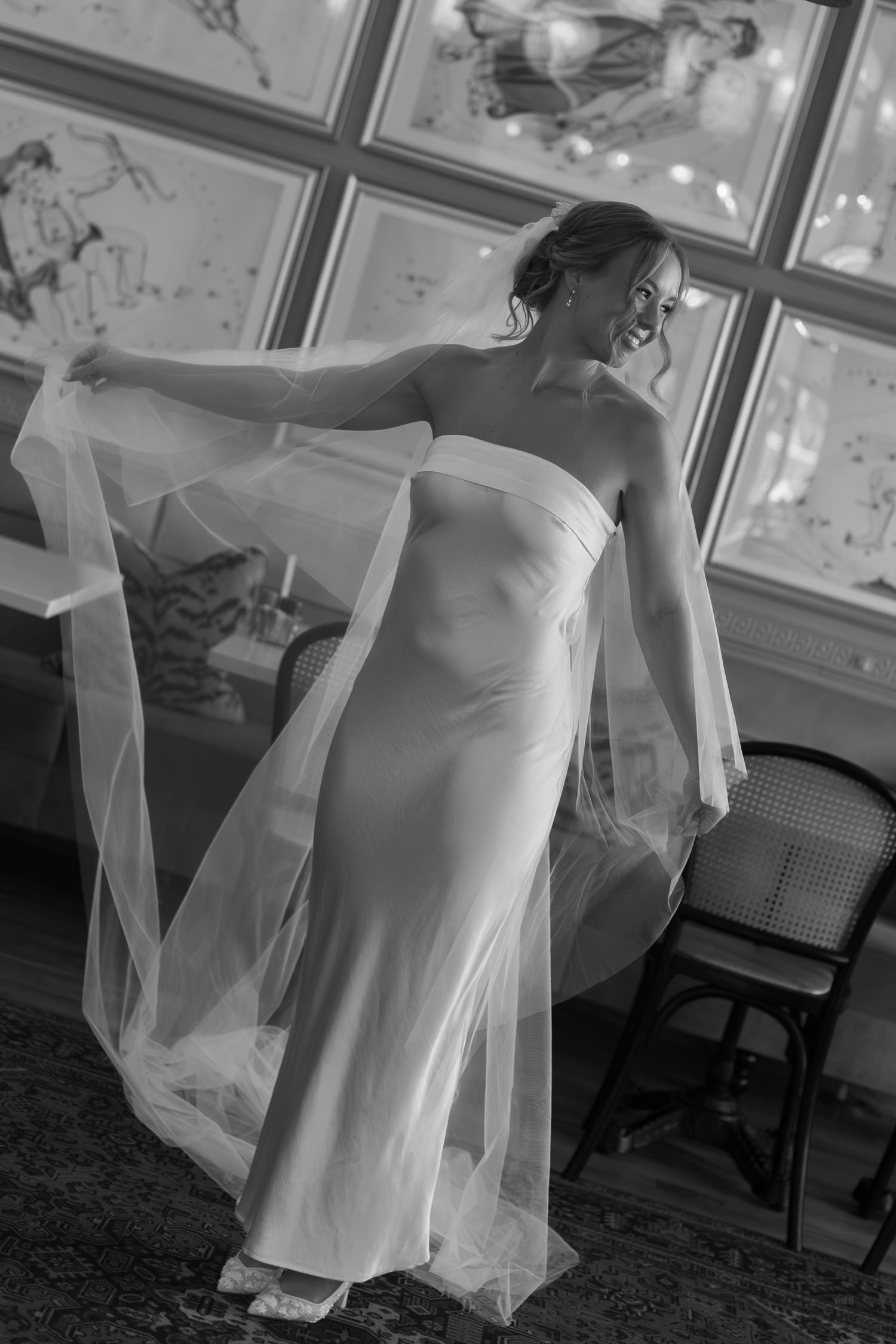 Artistic black and white photo of bride in flowing gown against framed wall decor.