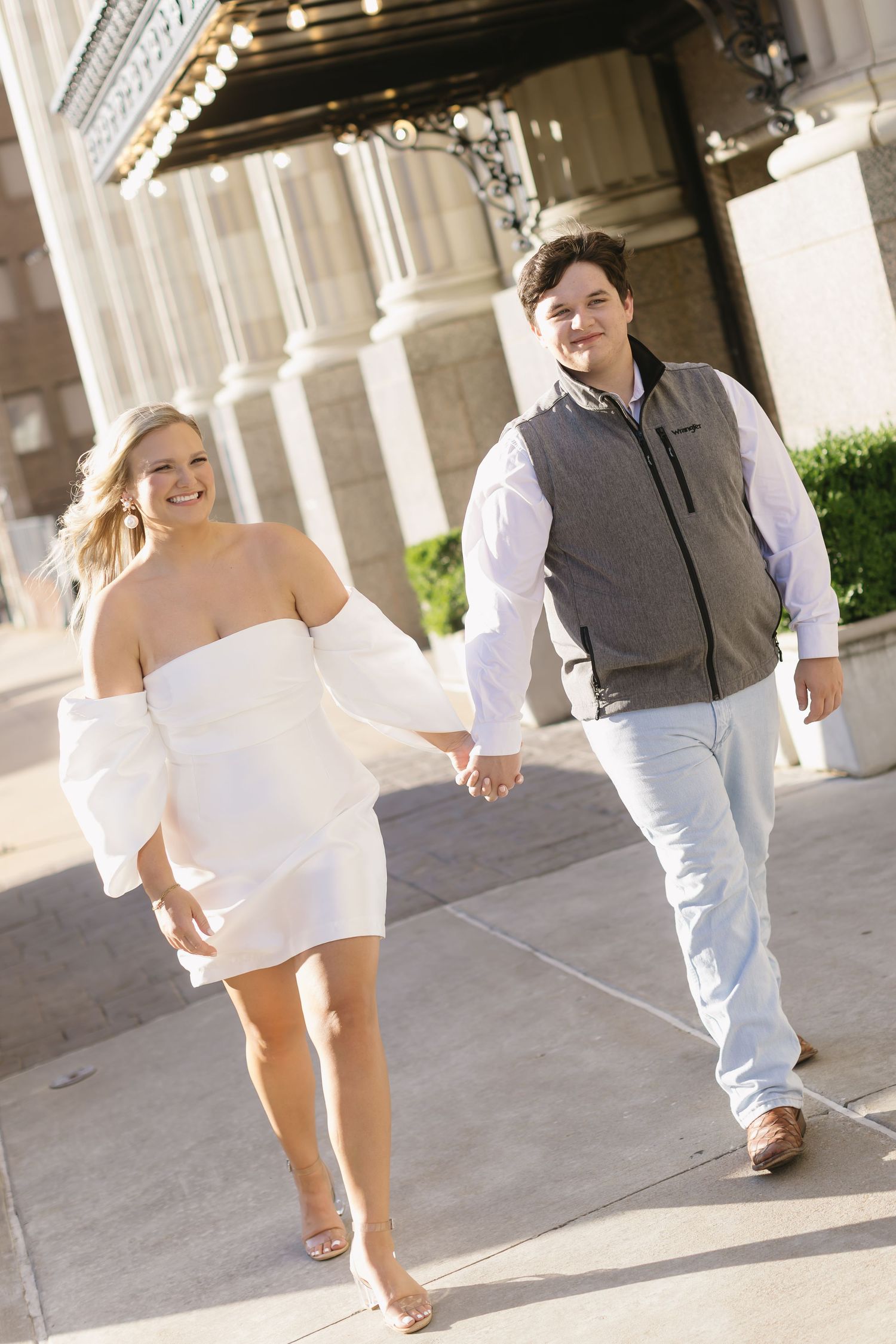 A couple holding hands and walking along a corridor with elegant columns and warm lighting.