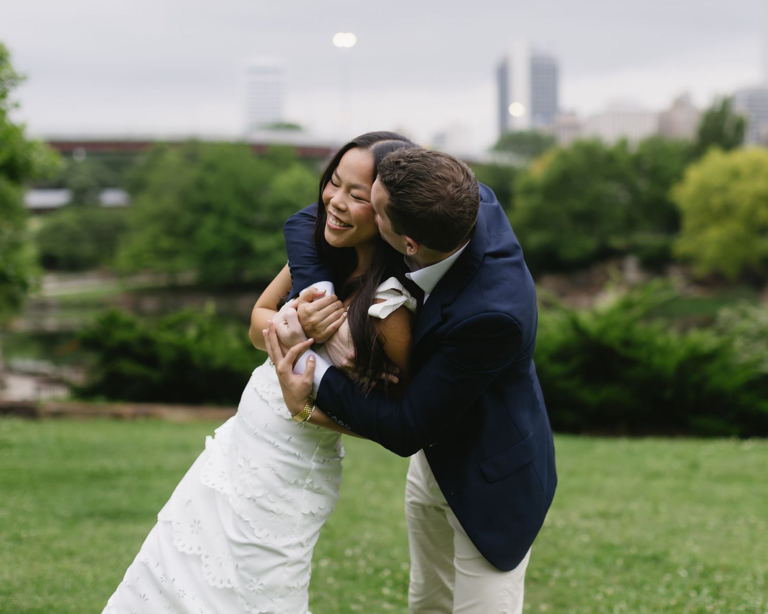 A couple in wedding attire share a playful dip on a green lawn surrounded by trees.