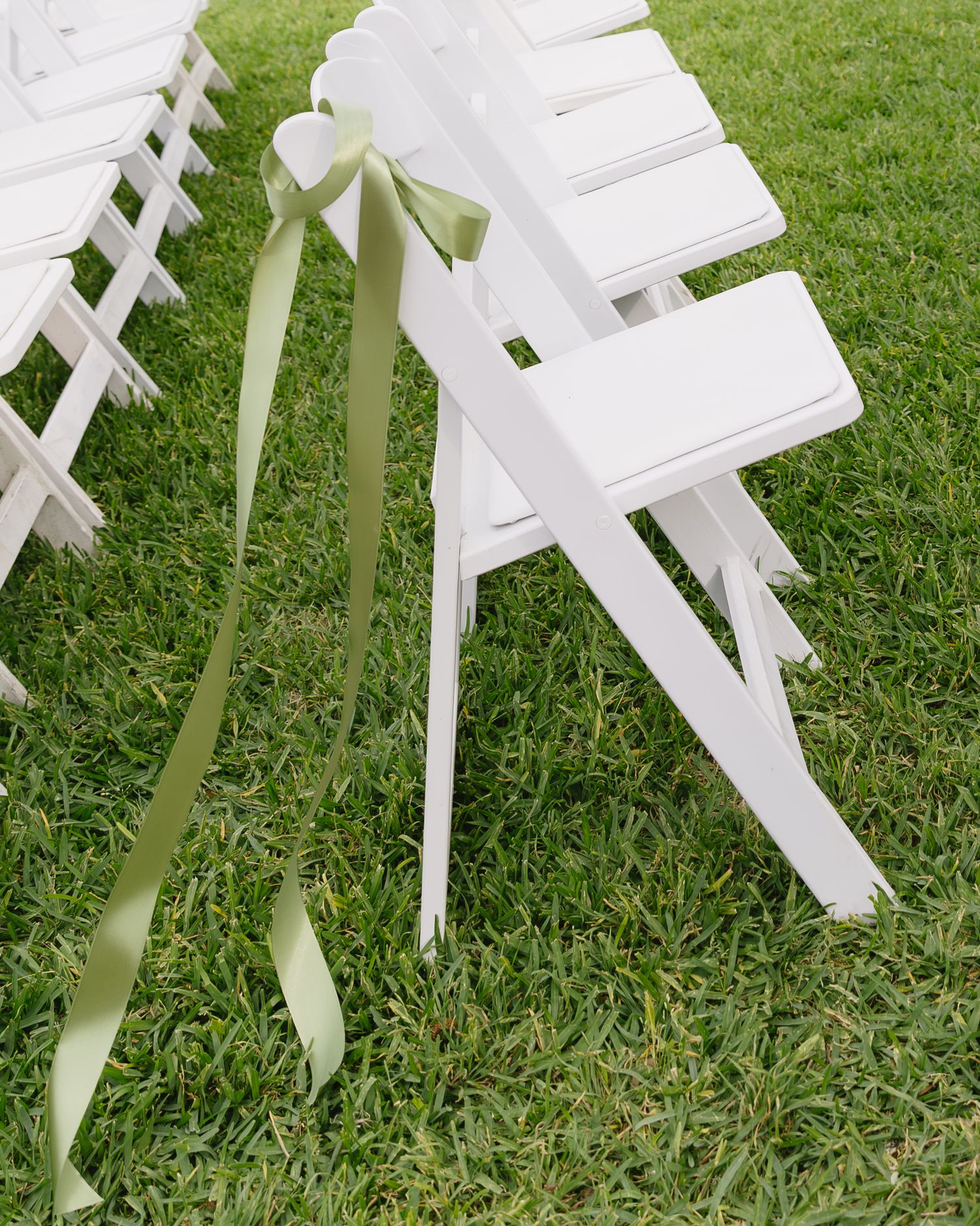 White folding chairs with sage green ribbons set up on grass for an outdoor wedding ceremony.