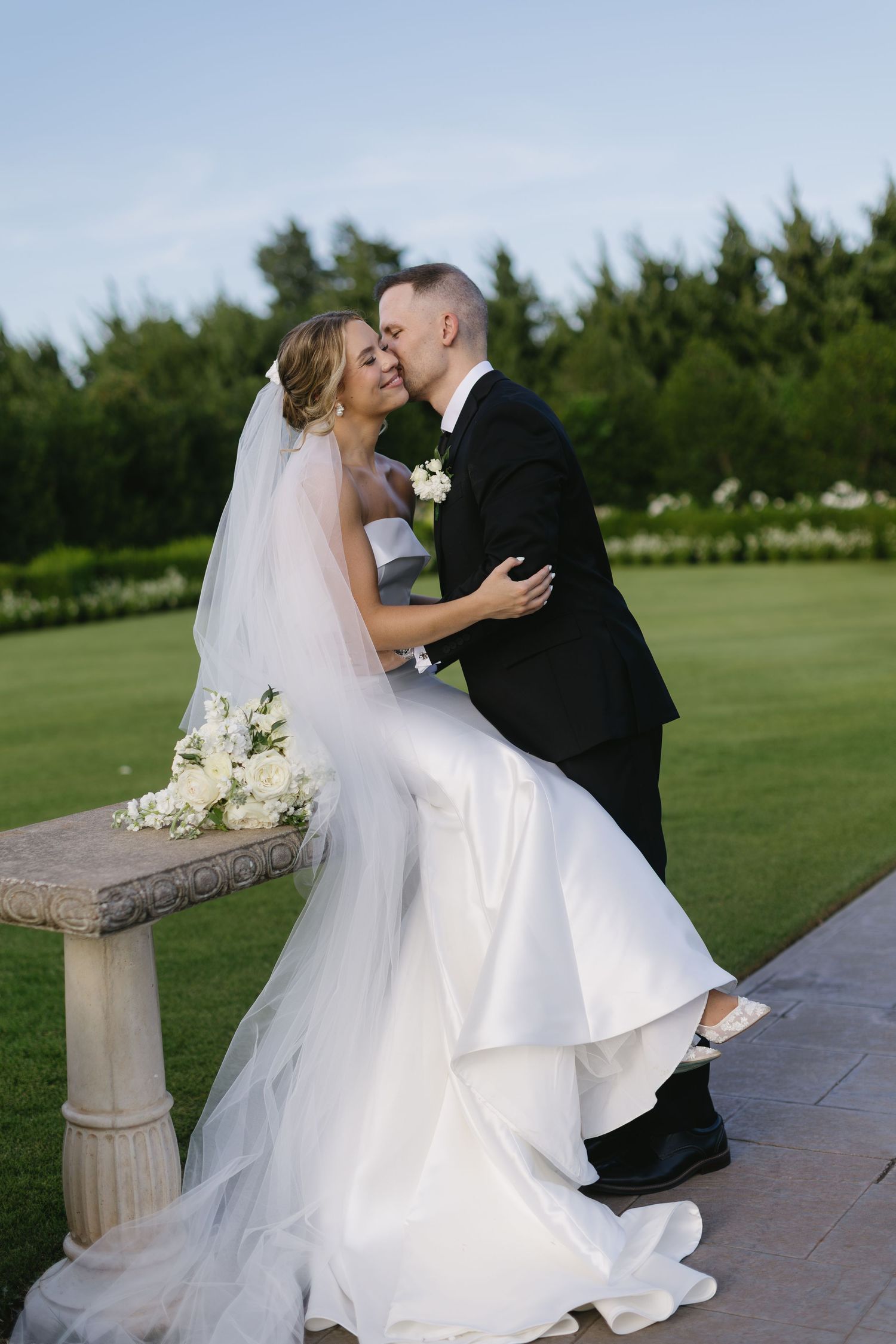 A romantic wedding moment captured outdoors with the bride in a flowing white gown seated on a stone bench.