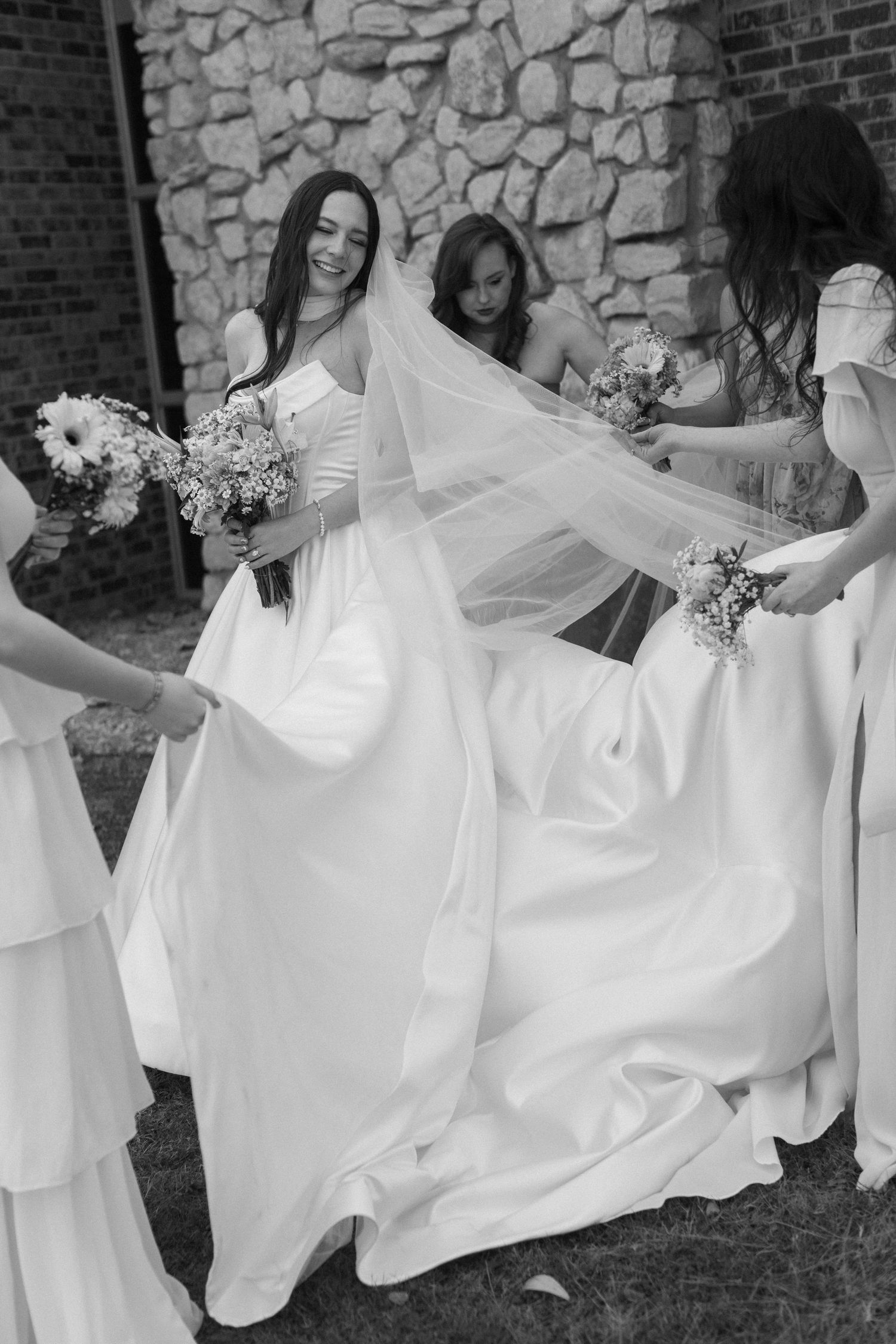 Bridesmaids in white dresses help arrange the bride's flowing gown and veil against a stone wall backdrop.