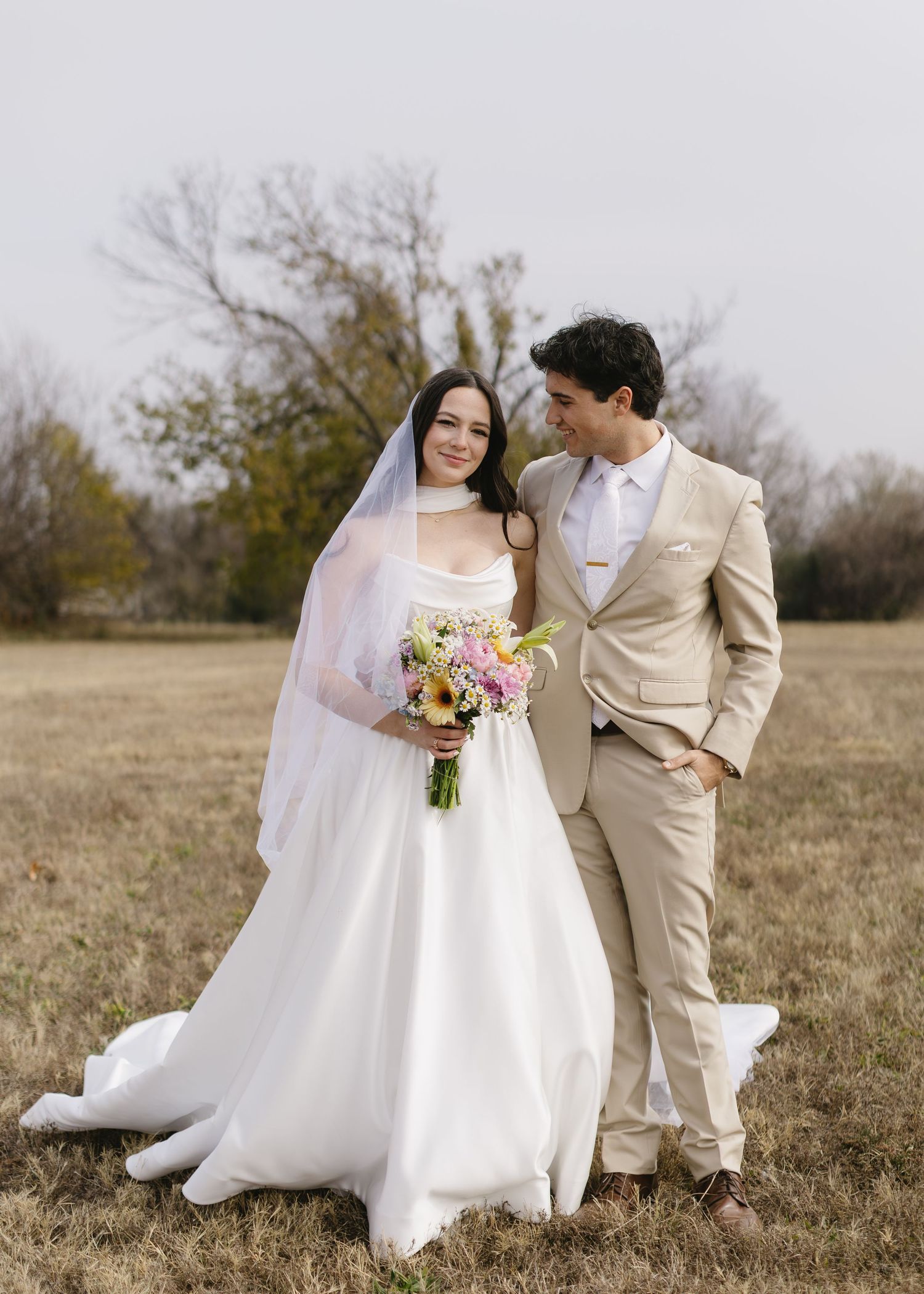 A couple shares intimate moments in a rustic field setting during their wedding photoshoot, wearing white and beige attire.