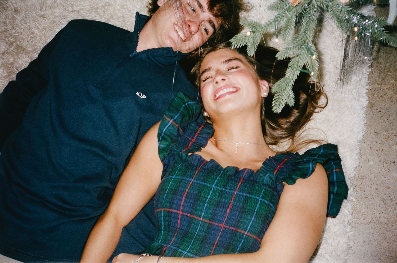 Two people lying down and laughing together near evergreen holiday decorations.