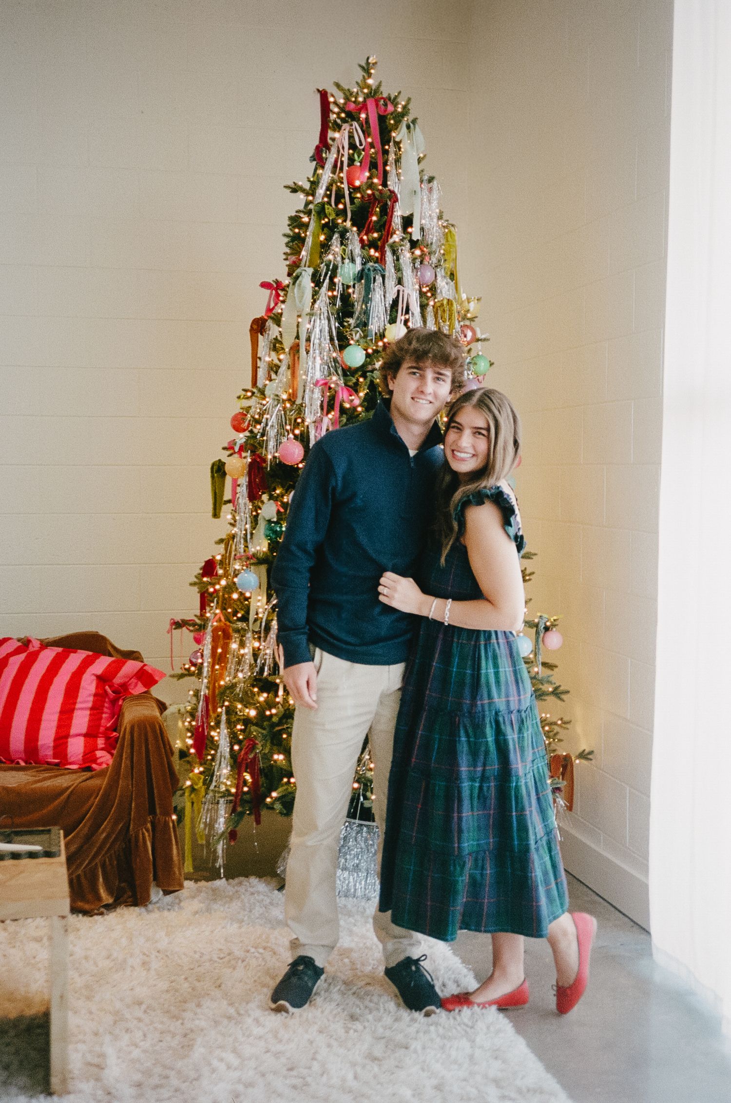 A couple poses in front of a decorated Christmas tree in a room with white walls and a red striped pillow.