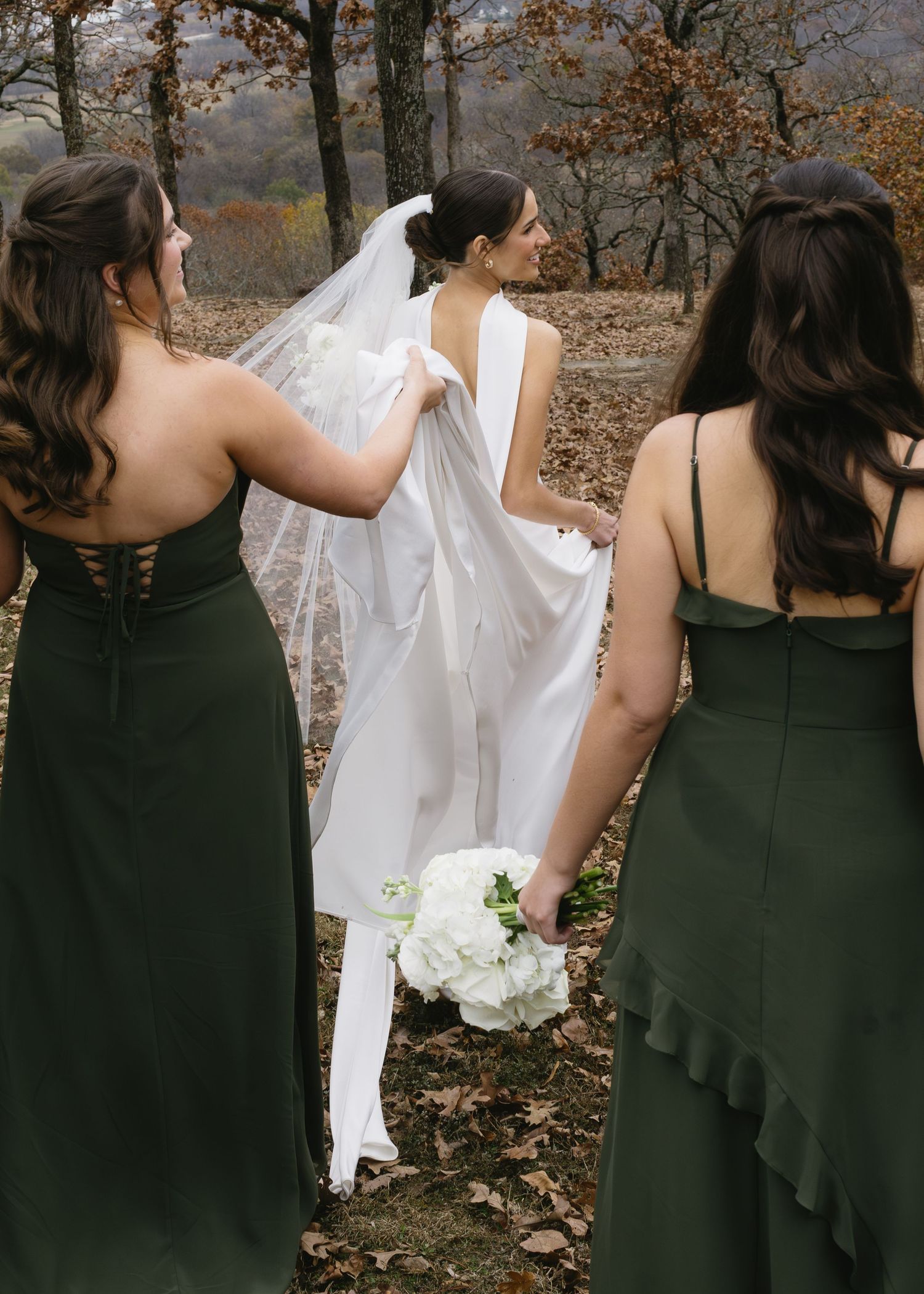 Bridesmaids in elegant dark green dresses help adjust the bride's veil in a rustic outdoor setting.