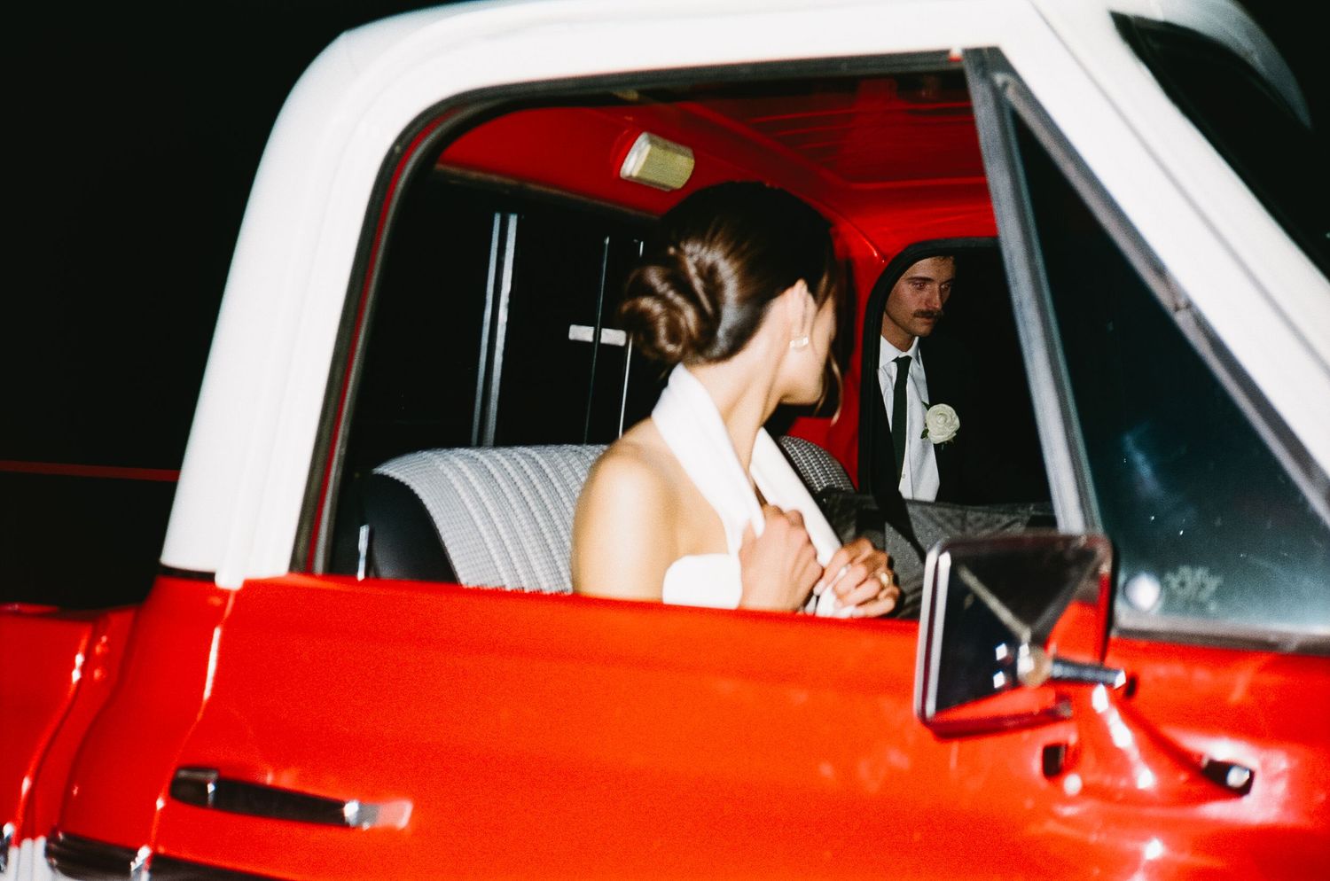 A newlywed couple sits in a bright red vintage car after their wedding ceremony.
