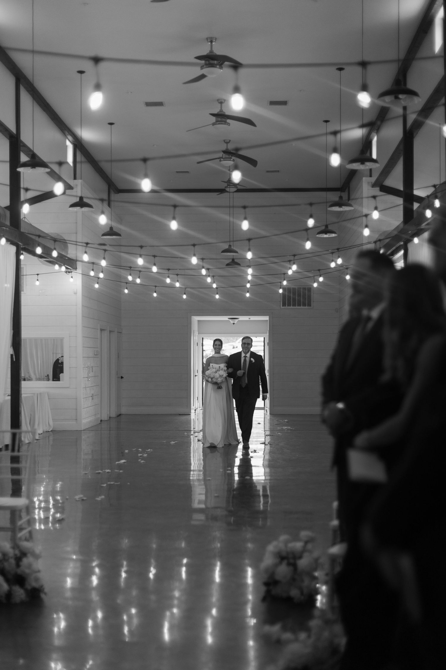 A dramatic black and white photo of a wedding venue with string lights and silhouettes in the distance.