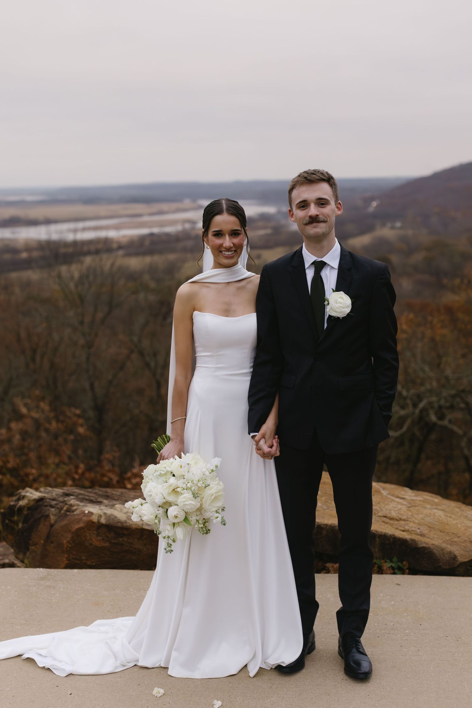 A wedding couple stands on an overlook with autumn scenery and rolling hills in the background.