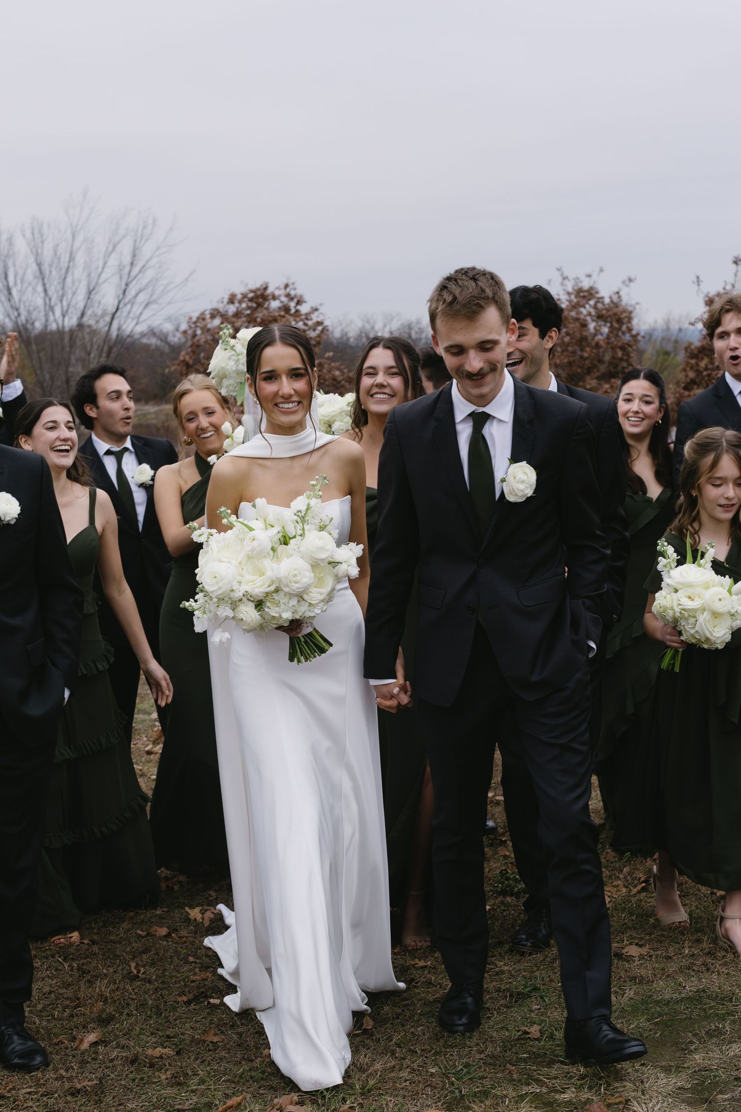 Wedding party in formal black attire walks together through autumn landscape.