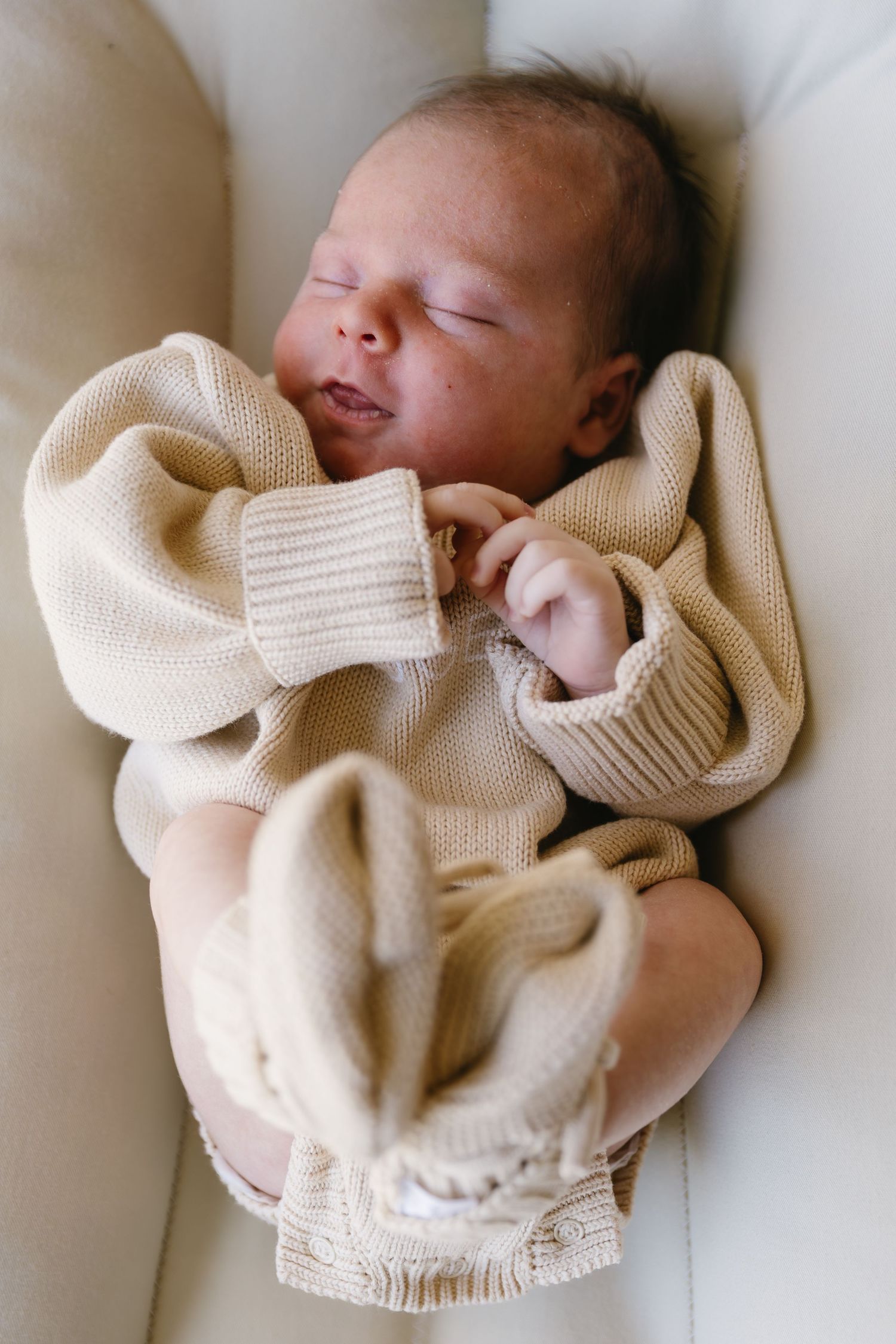 A sleeping newborn baby wrapped in a cozy cream-colored knit sweater lies peacefully on white bedding.