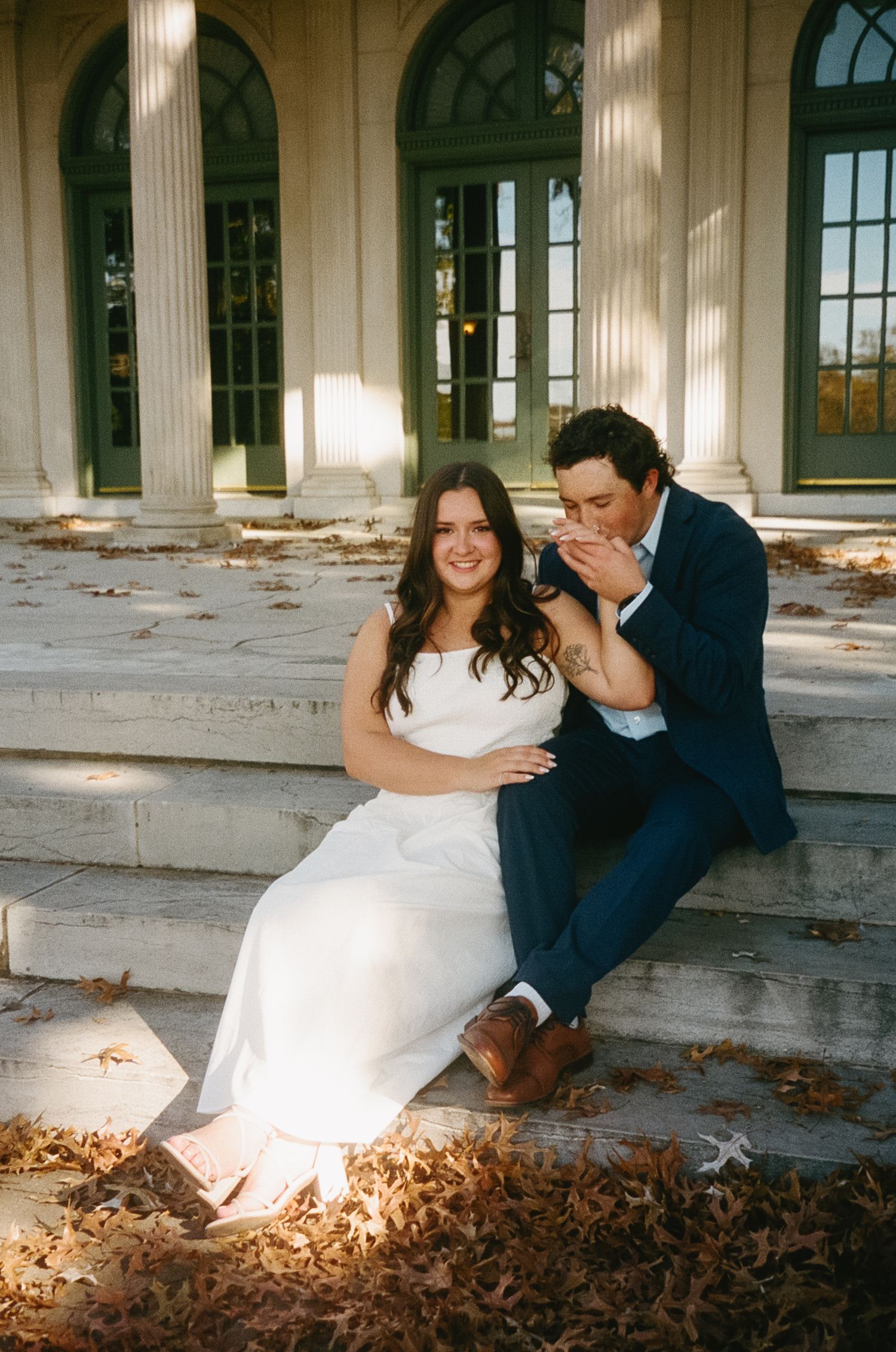 A couple shares playful moments on stone steps surrounded by autumn leaves at a historic columned building.