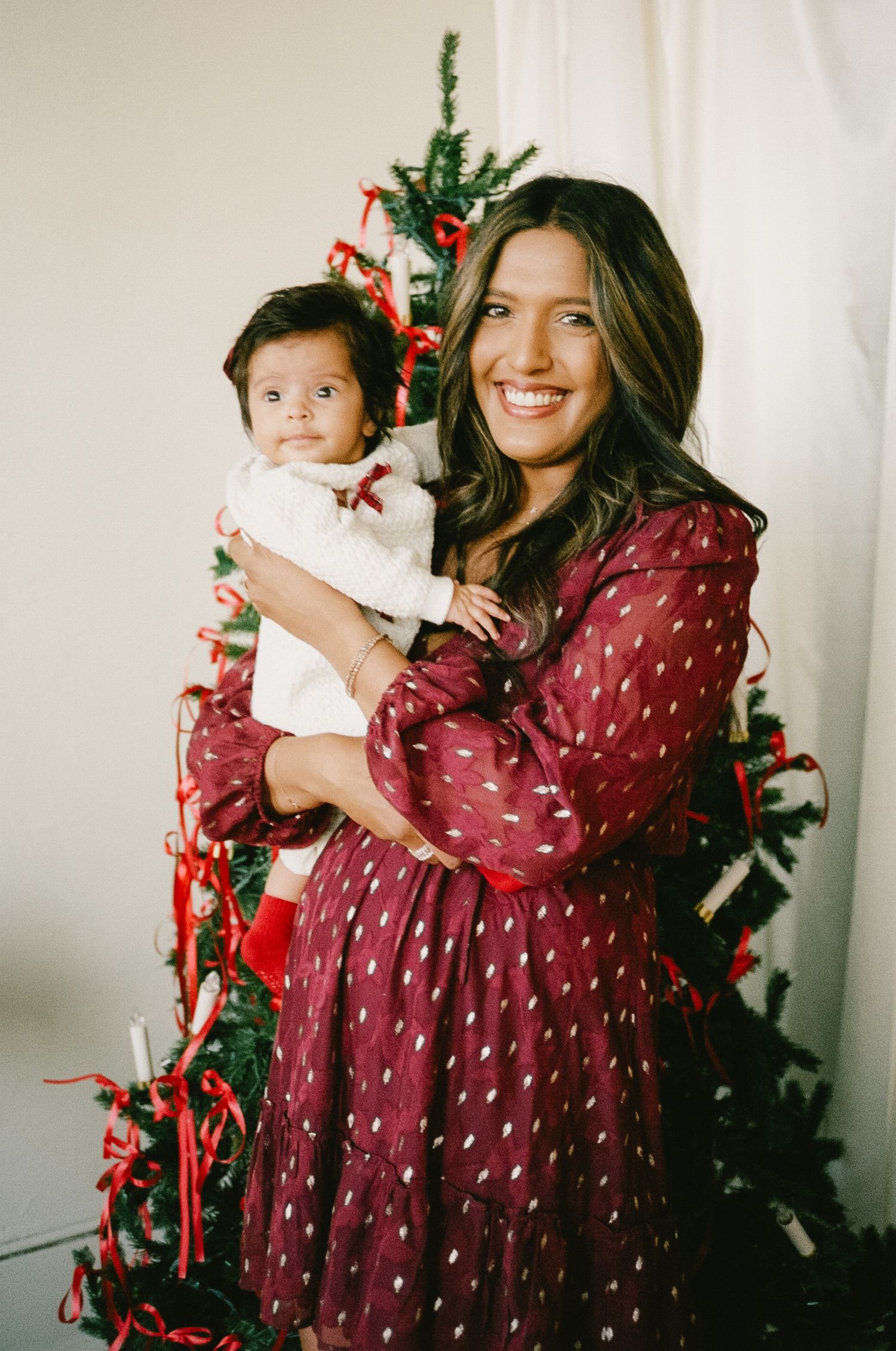 A person in a burgundy polka dot dress poses playfully in front of a Christmas tree in a photo series.