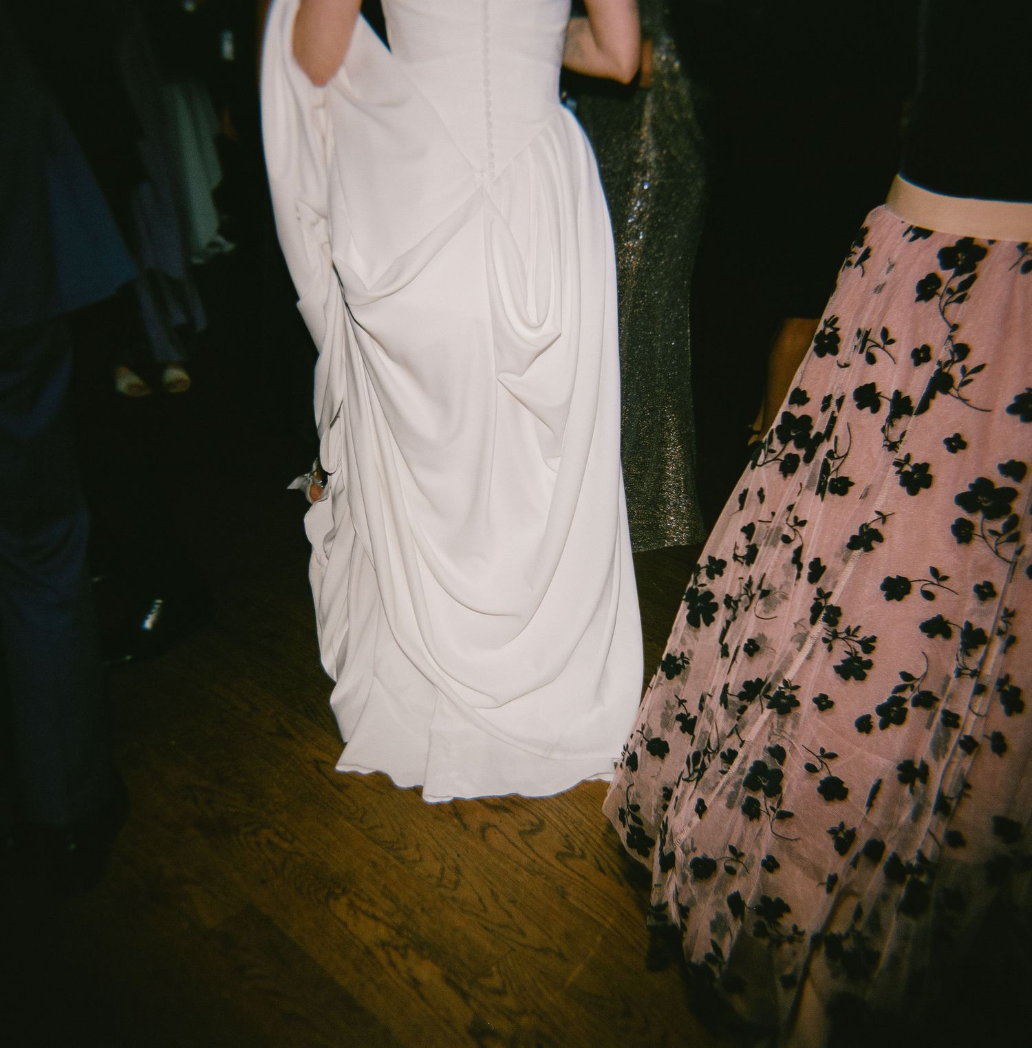 A white wedding dress and pink floral dress are captured on a wooden dance floor at a celebration.