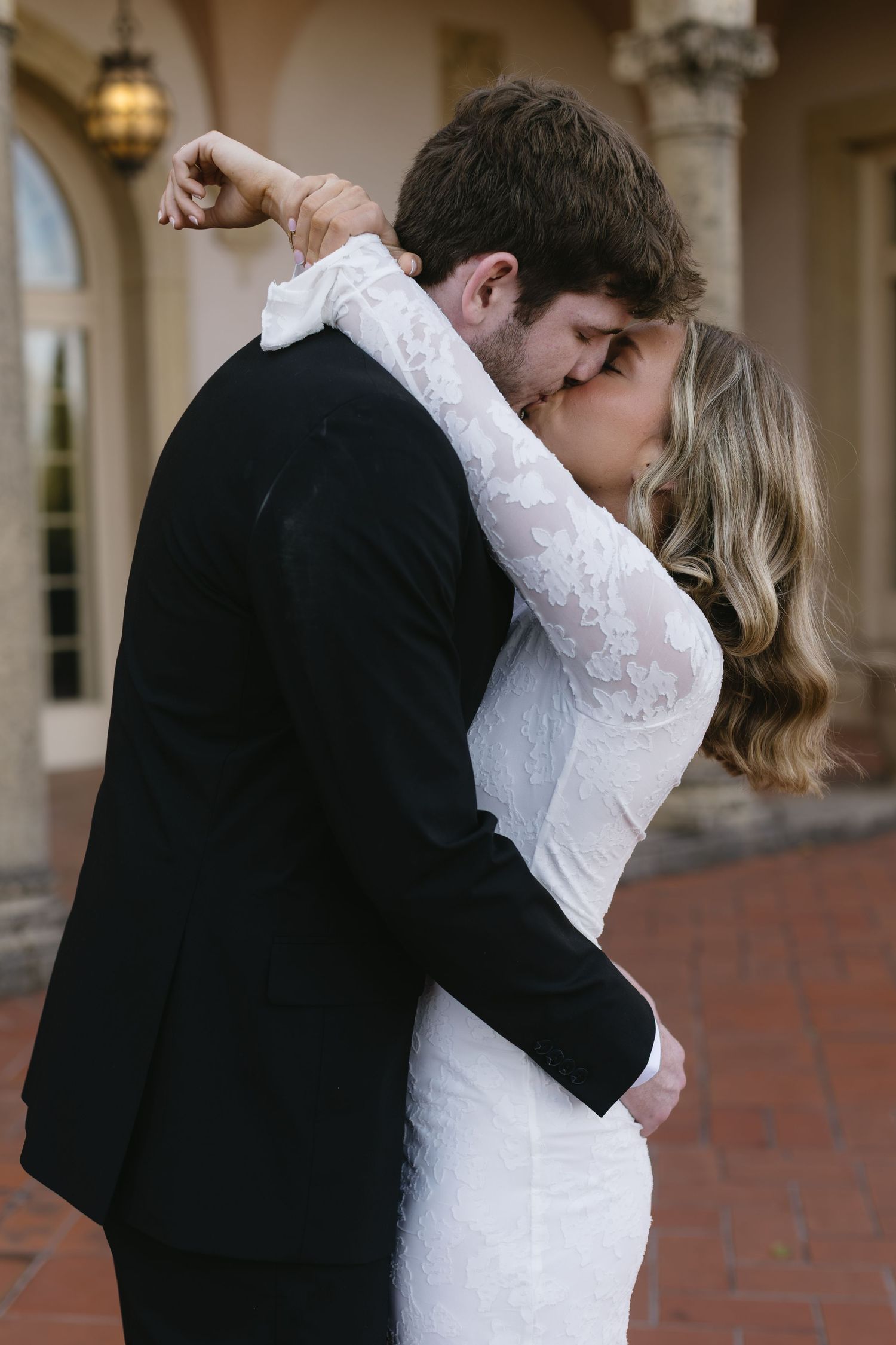 A couple embraces intimately in an elegant courtyard setting with stone columns and brick flooring.