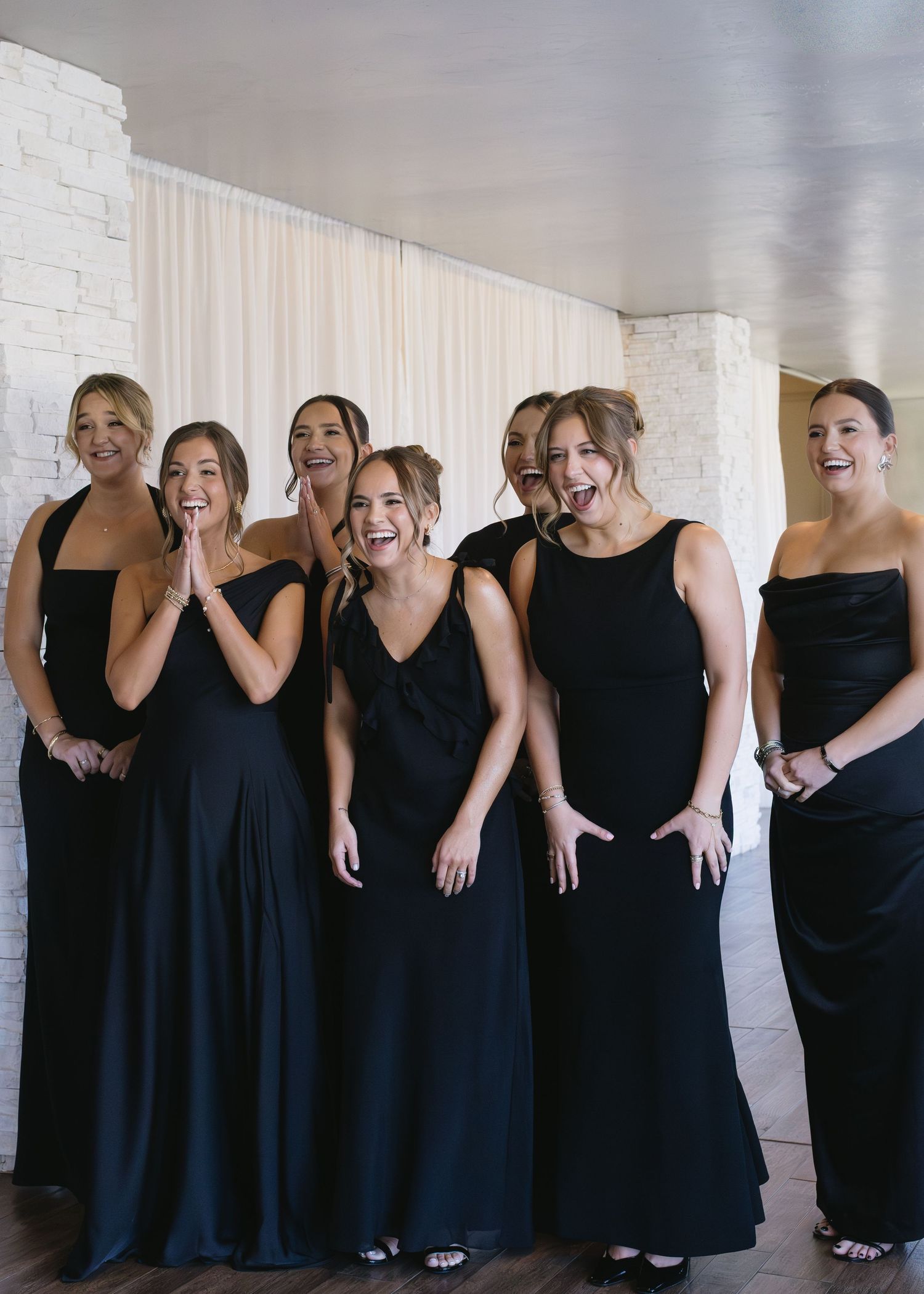 Bridesmaids in matching black dresses laughing together against a white brick wall.