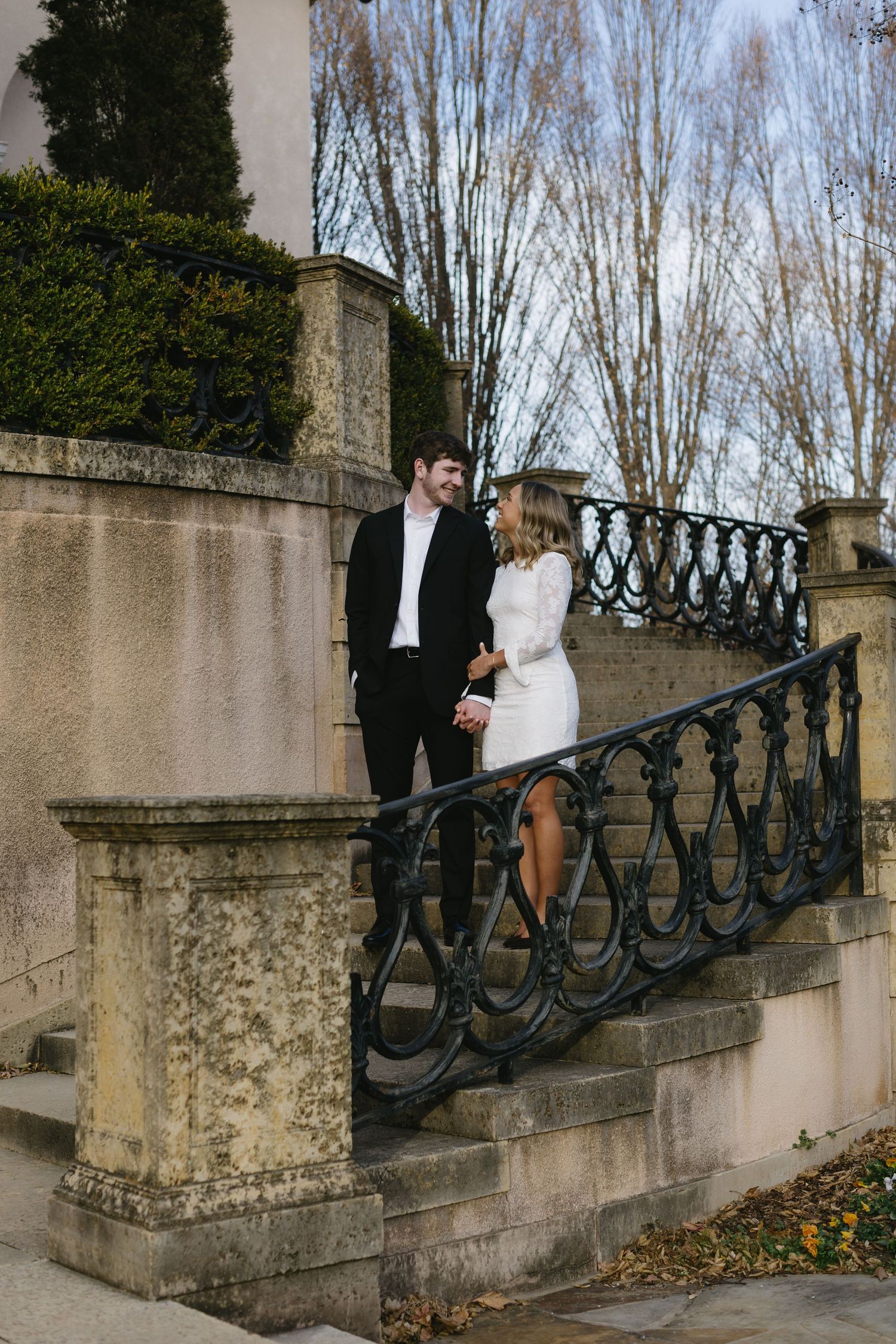 A couple stands on stone steps with ornate black metal railings against a winter backdrop.