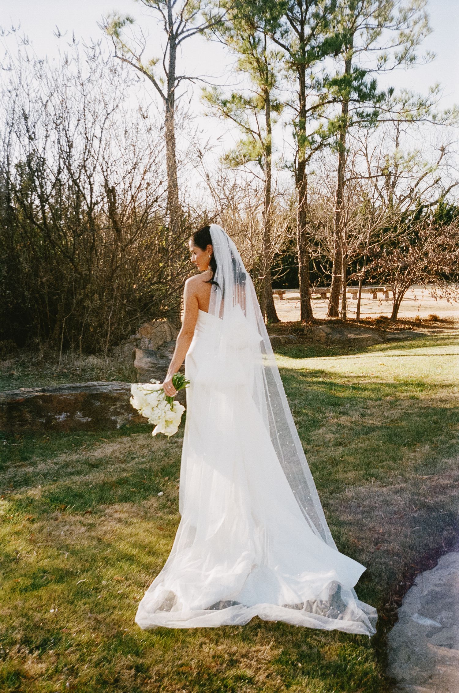 Bride in flowing white dress and veil poses in outdoor garden setting.