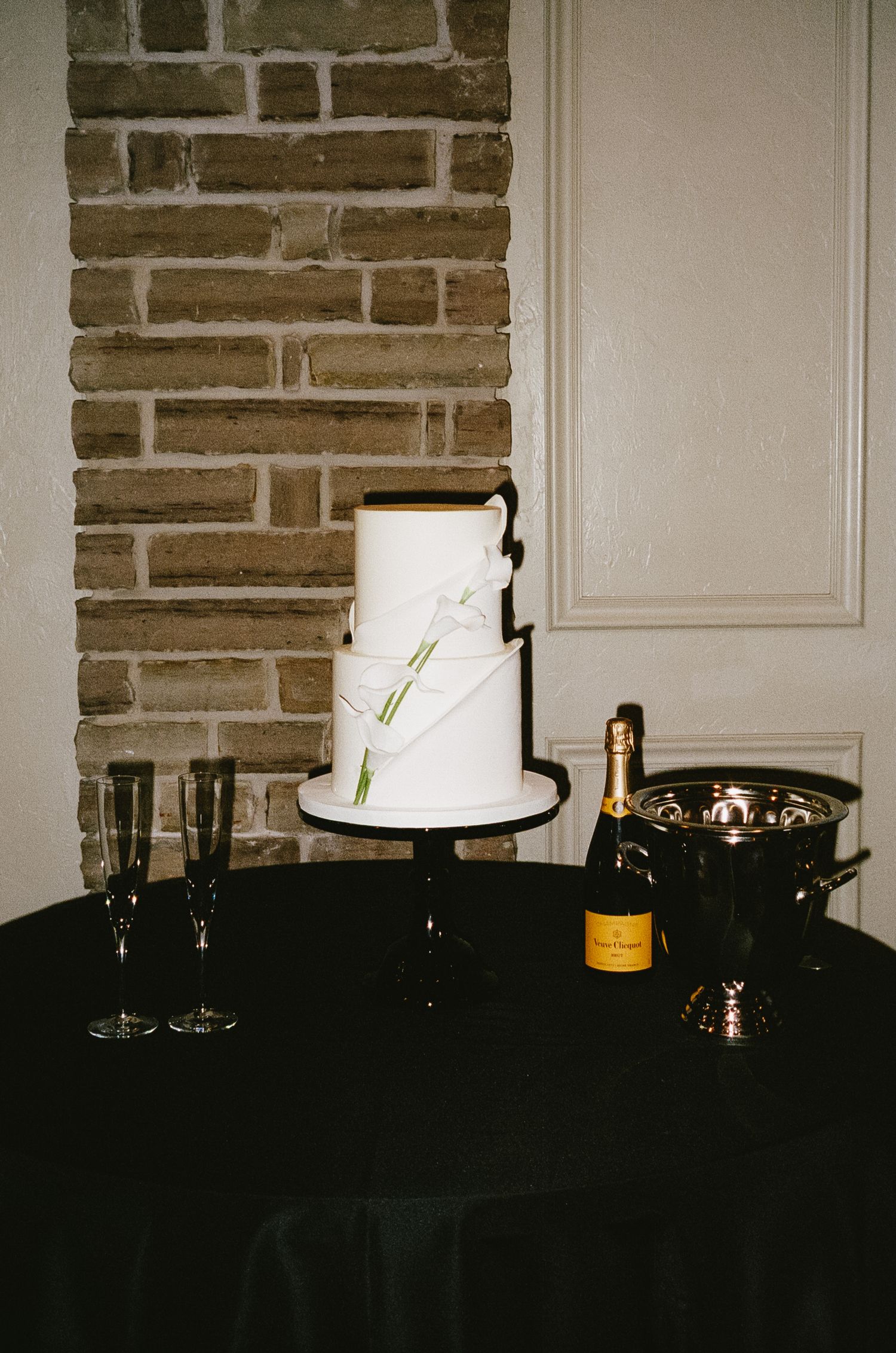 A white wedding cake with simple floral accents sits on a dark table against an exposed brick wall.