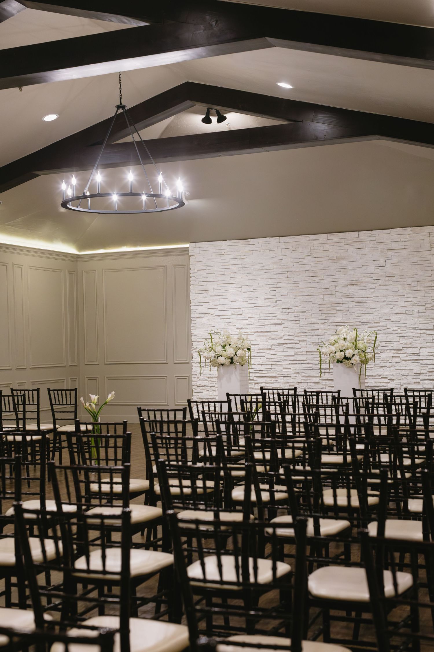 Modern wedding ceremony space with black chiavari chairs and circular chandelier against white stone wall.