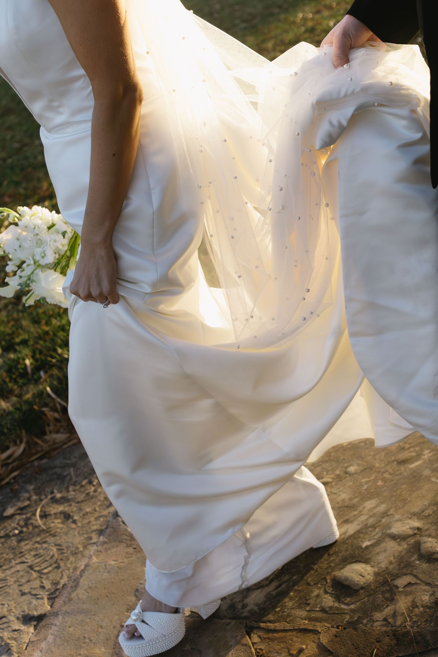 Close-up details of a white wedding dress hem and shoes on a stone path.