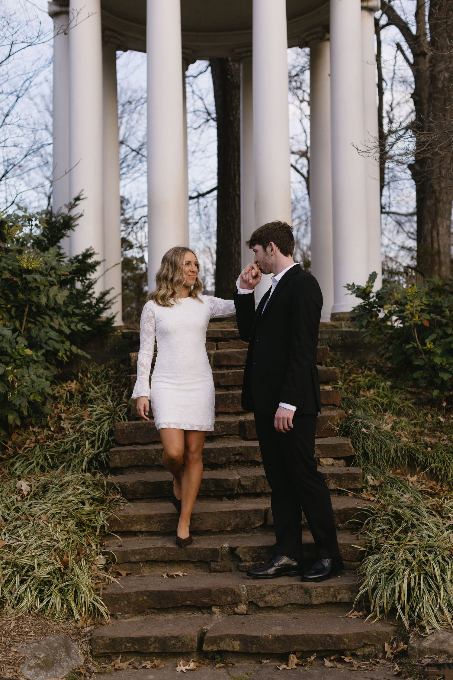 A couple poses on stone steps beneath classical white columns surrounded by winter foliage.