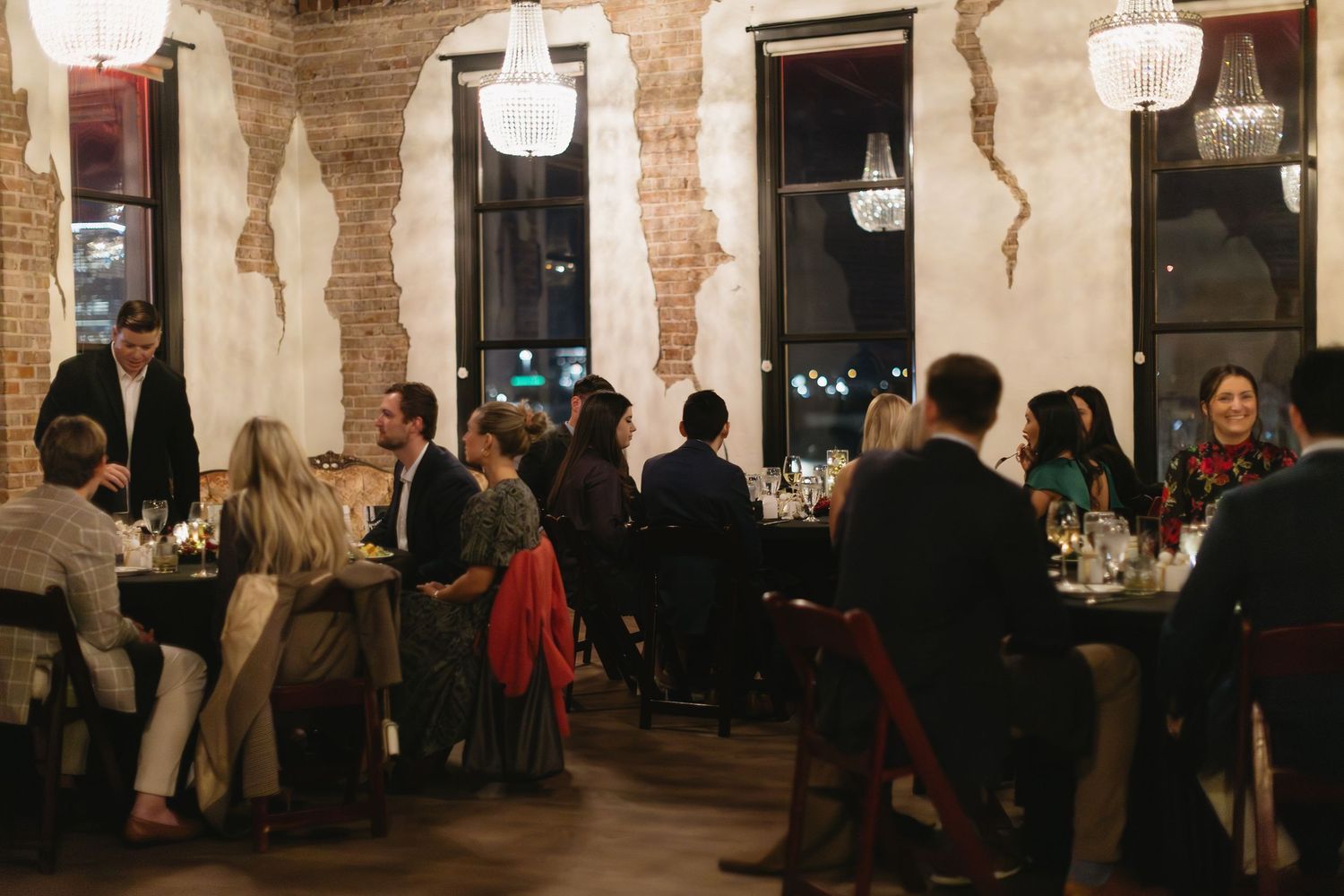 Guests gather at dining tables in a rustic venue with exposed brick walls and pendant lighting.