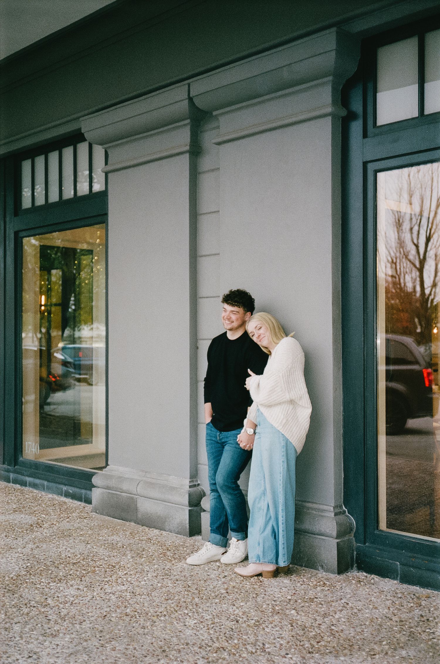 A couple poses playfully against a white building wall with dark trim.