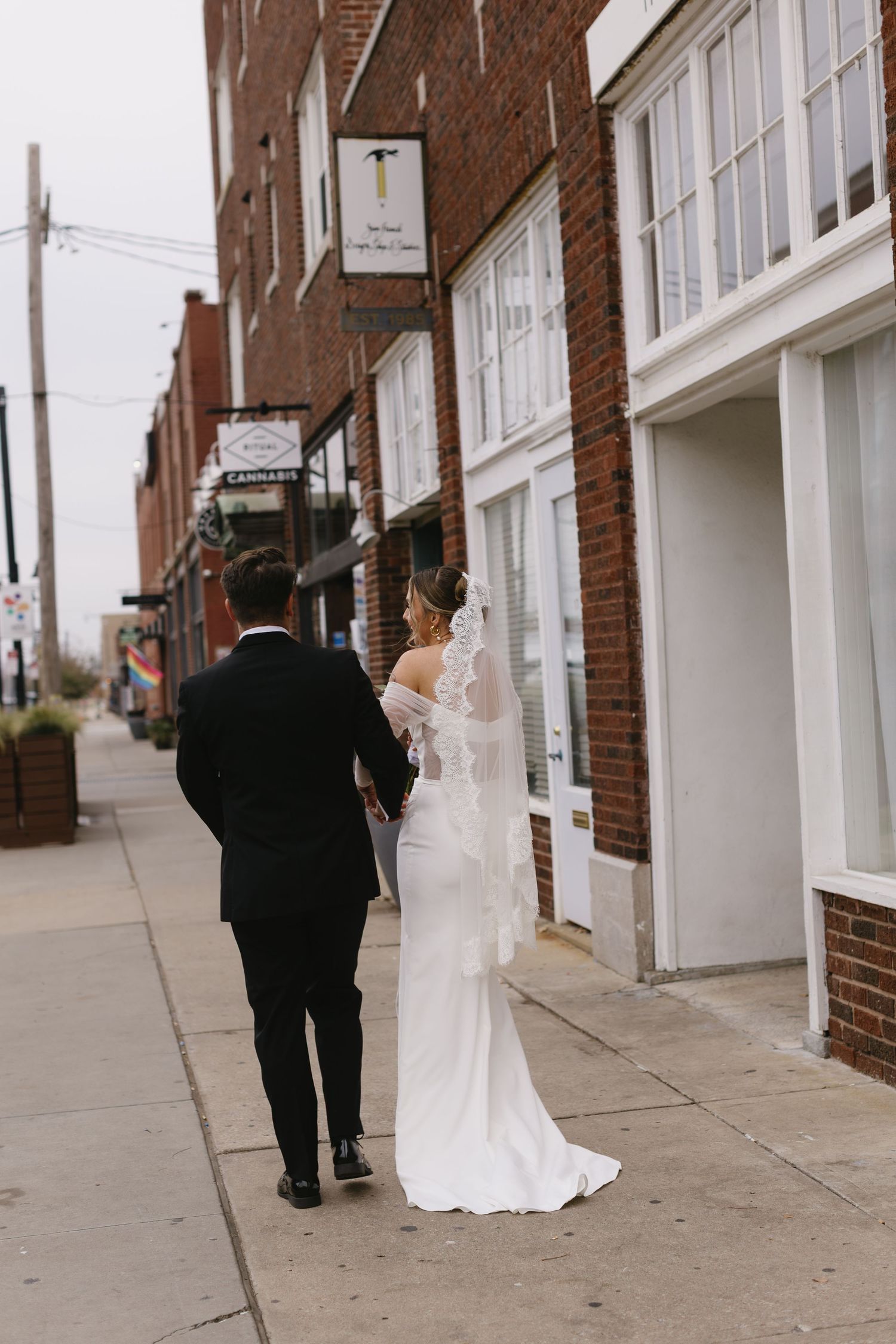 A couple in formal wedding attire walks along a brick building sidewalk.